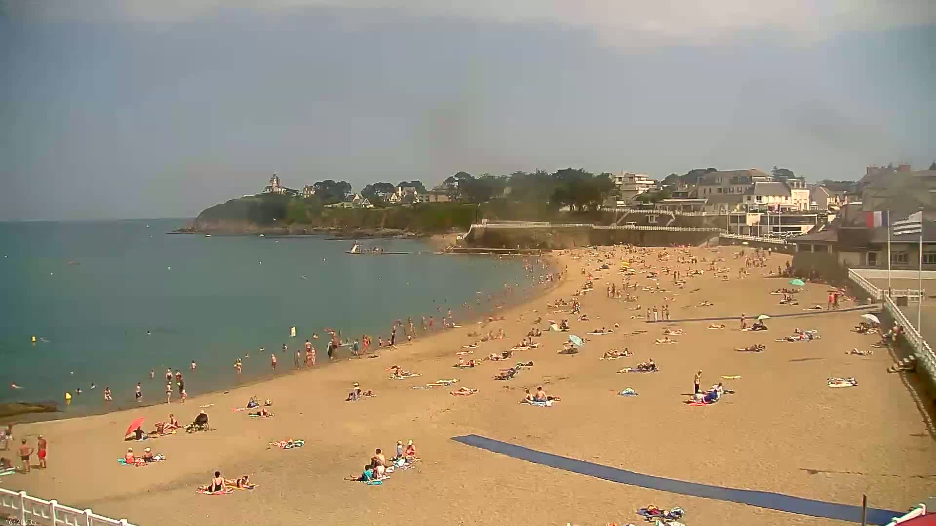 A sandy beach with many people sunbathing and swimming, backed by buildings and a calm ocean under a mostly sunny sky.