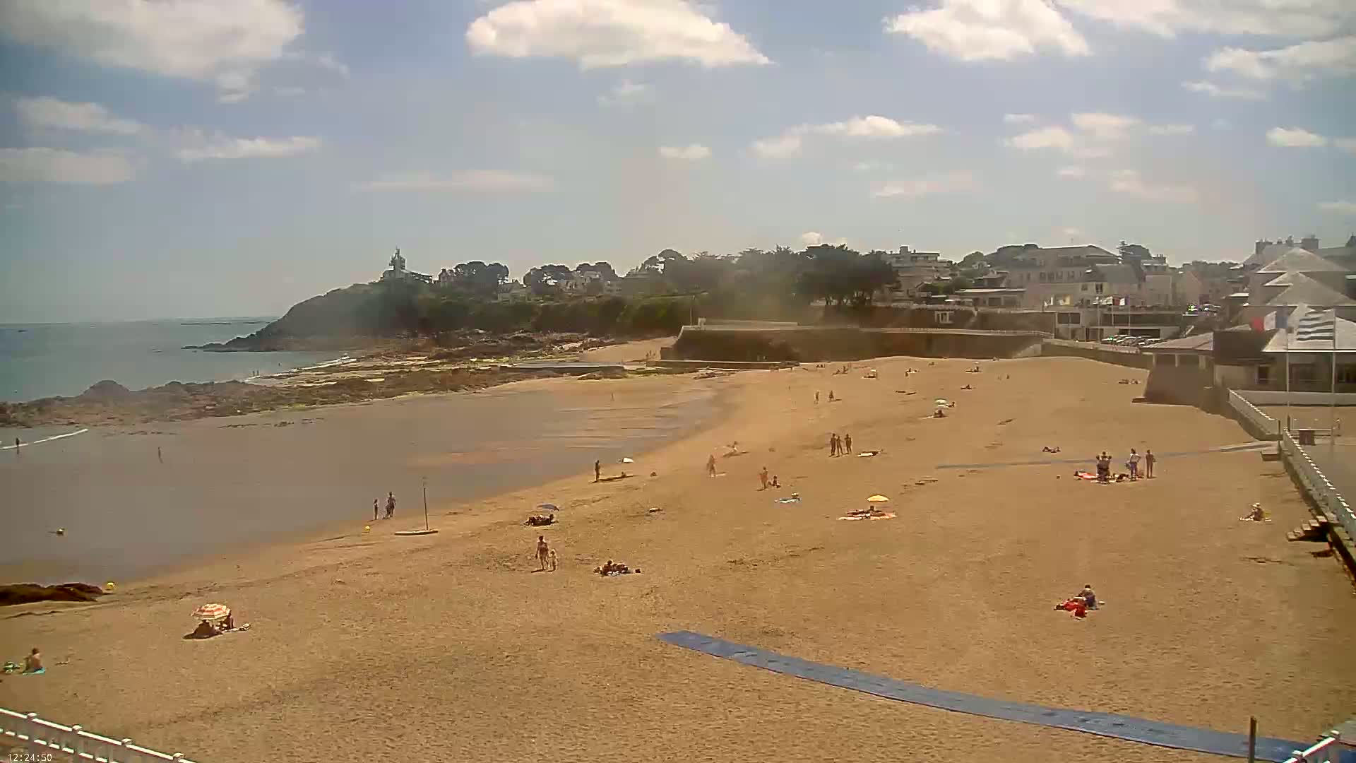 A sandy beach with scattered sunbathers stretches from calm ocean water to a seaside town under a partly cloudy sky.
