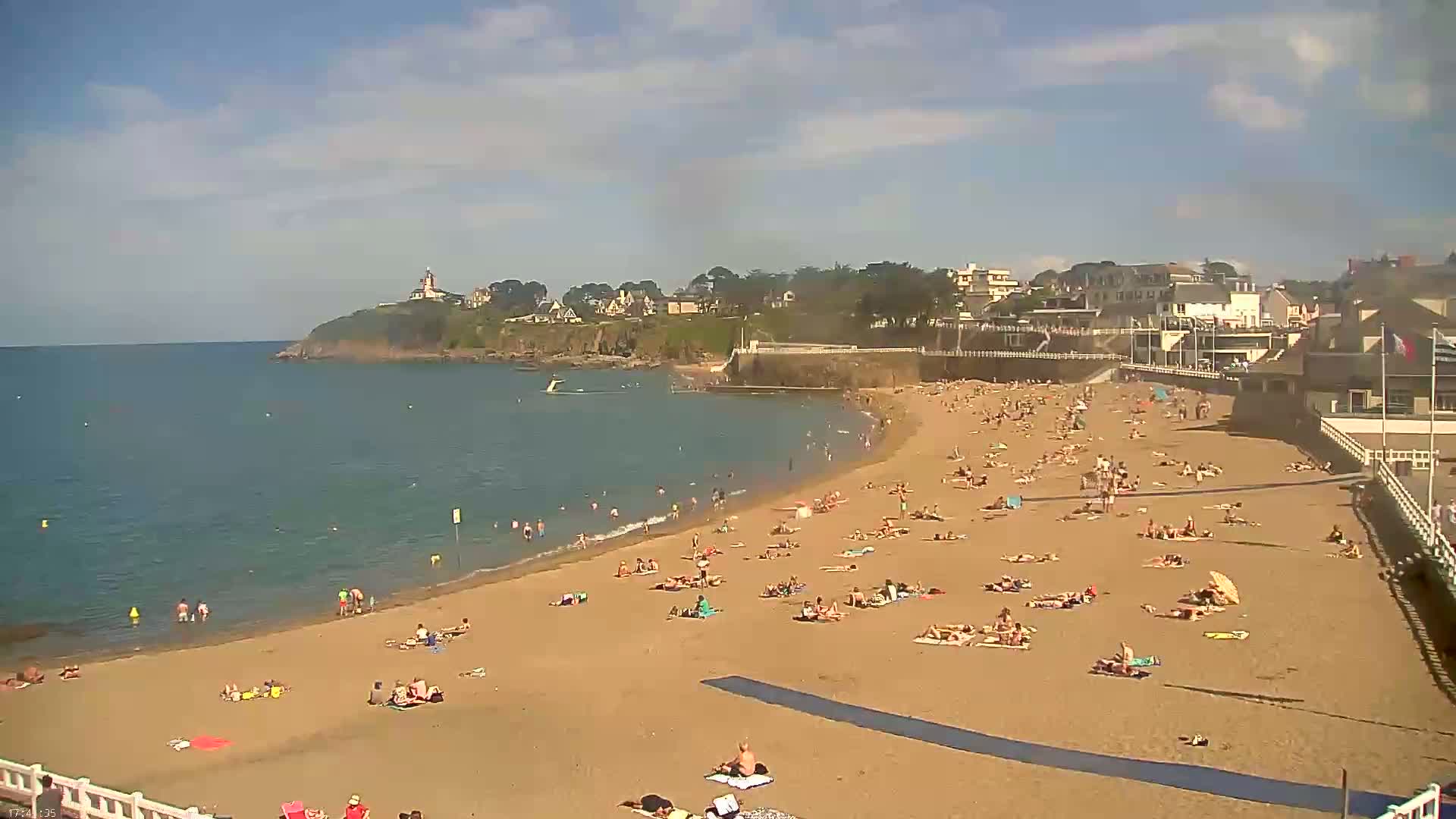 A sandy beach with many people sunbathing and swimming in calm, clear water, under a mostly sunny sky, is nestled in a bay next to a town with buildings and a lighthouse on a nearby headland.