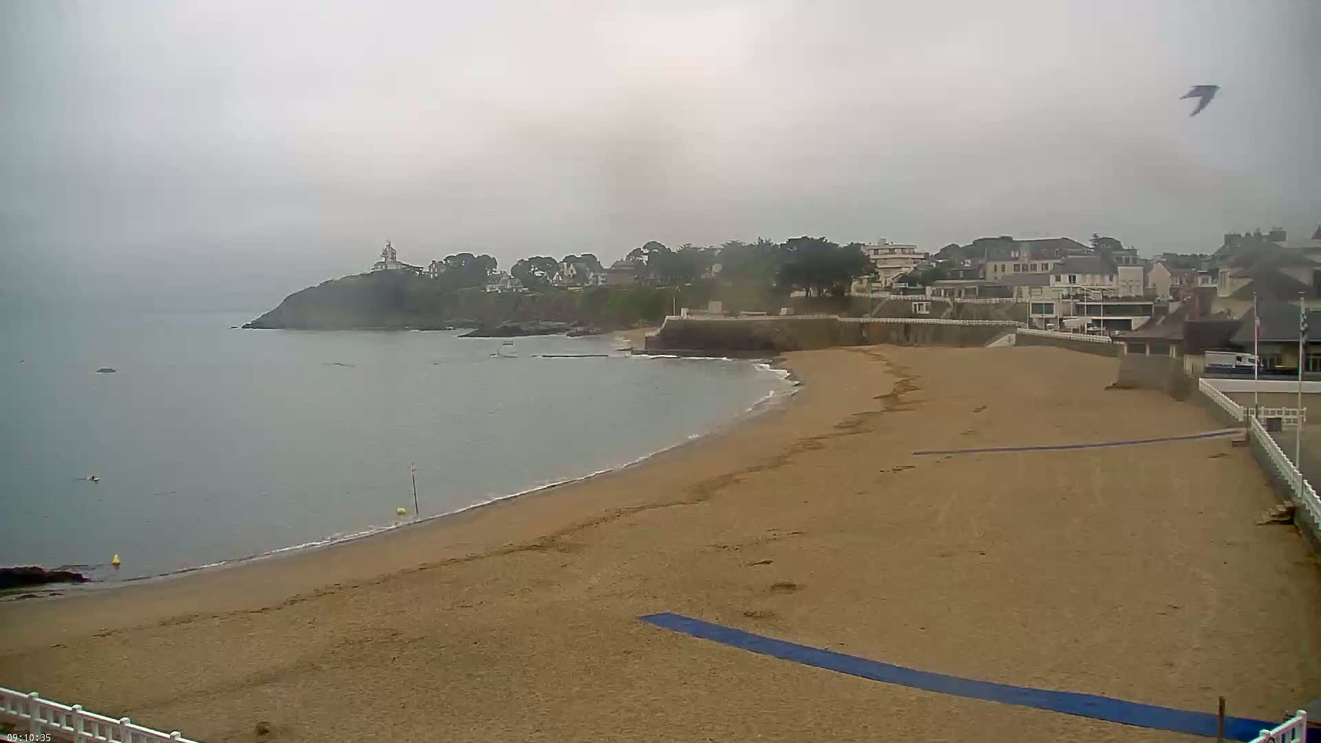 A sandy beach curves along a calm ocean under an overcast sky, with a small town visible on a hillside in the distance.