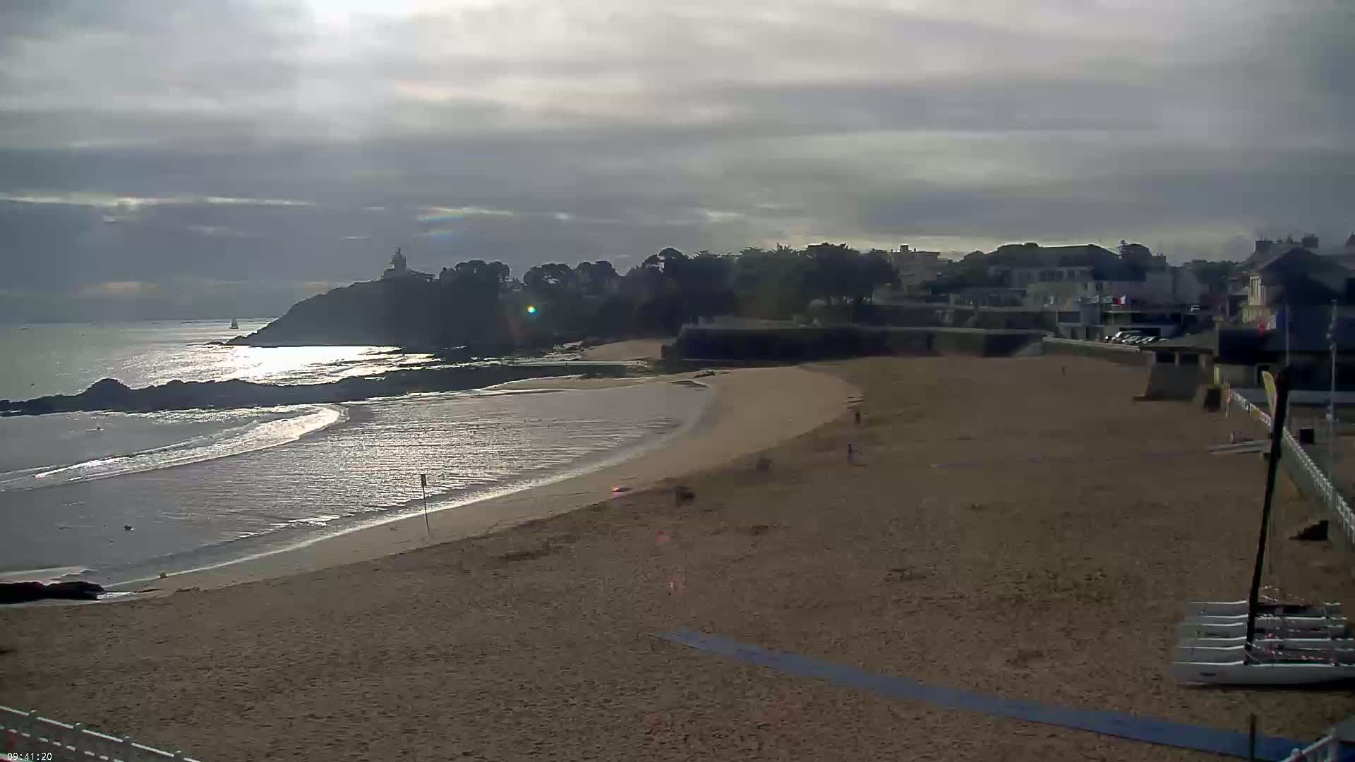 A sandy beach curves along a calm, sunlit ocean, backed by a line of buildings and green vegetation under a mostly cloudy sky.