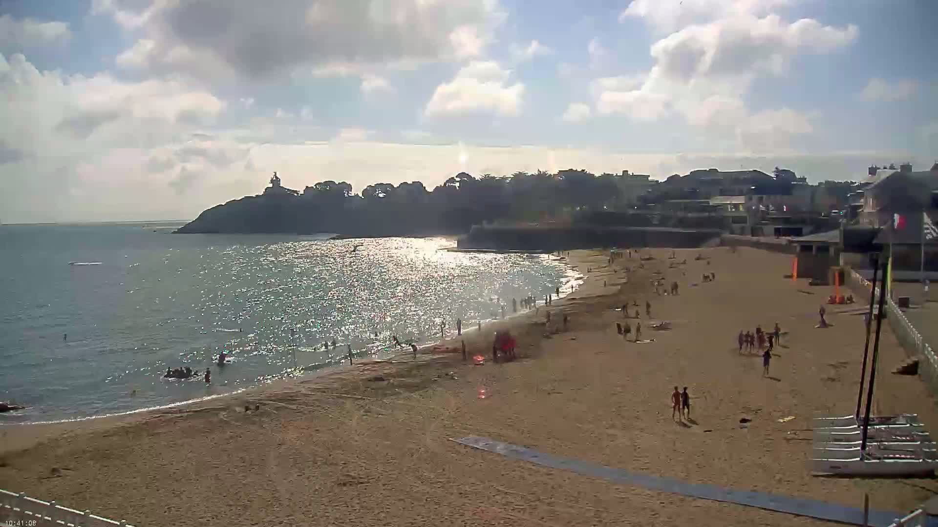 A sandy beach with many people on it and in the water, under a partly cloudy sky, is bordered by a calm sea and a line of buildings and trees.