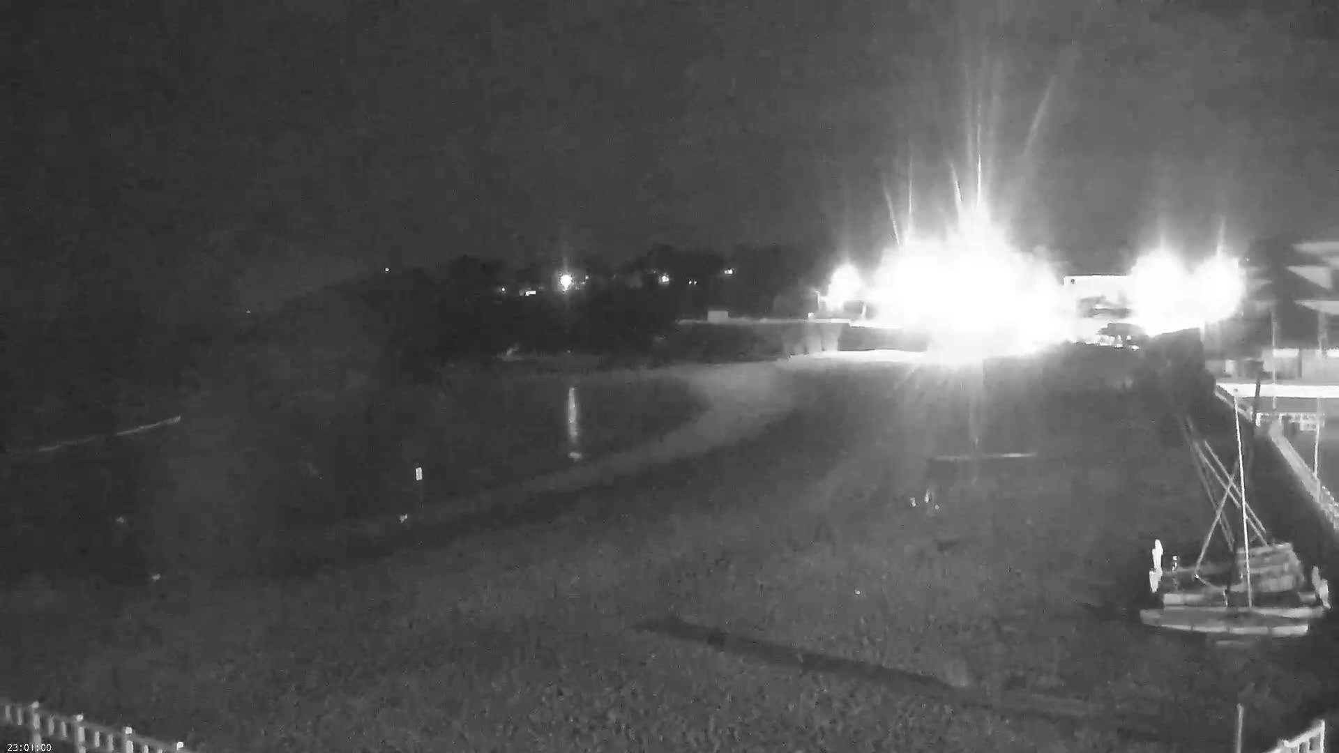A nighttime, low-light view shows a dark beach with some structures and boats in the foreground, with bright lights and dark vegetation visible in the background.