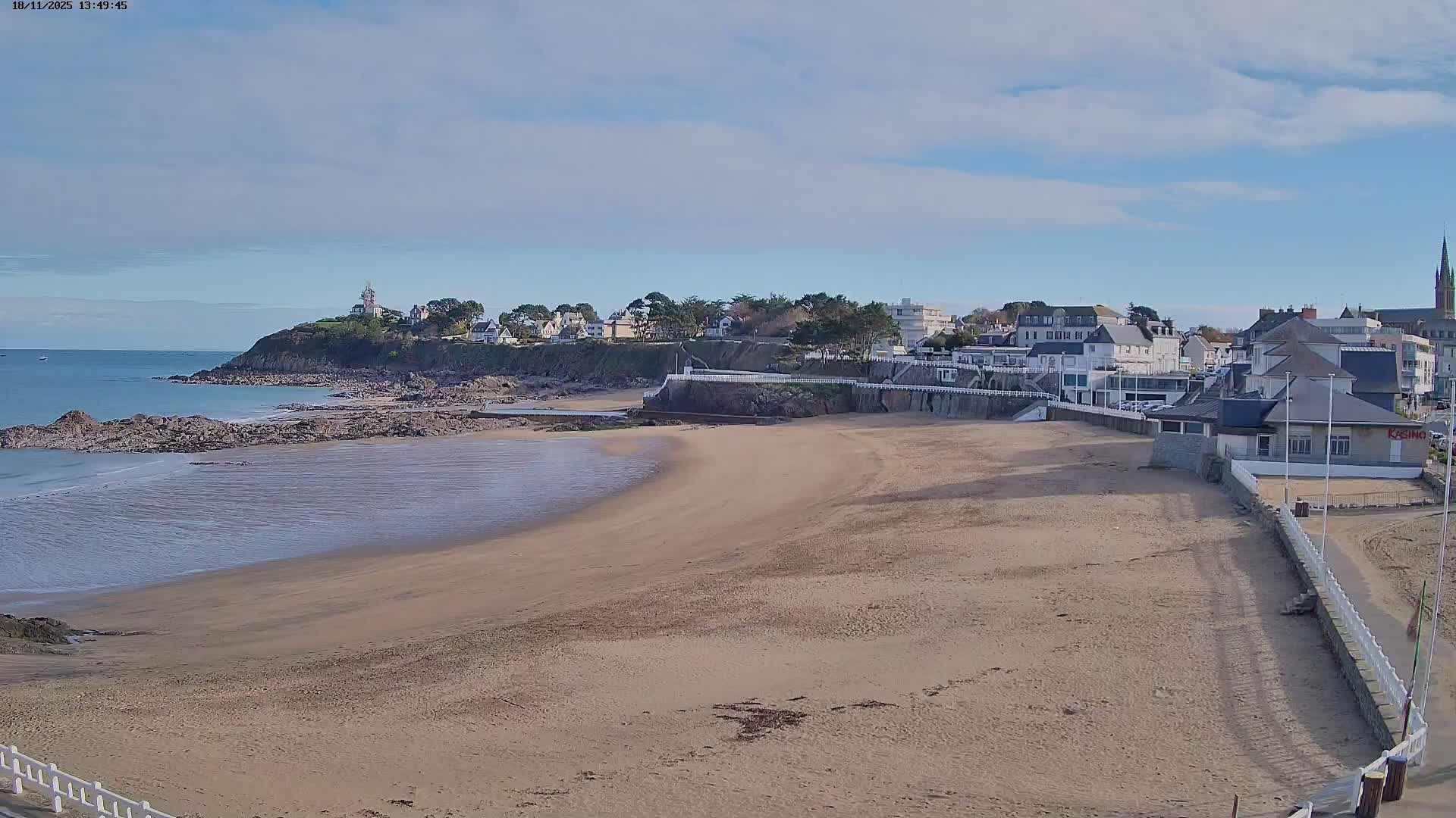 A nighttime, low-light view shows a dark beach with some structures and boats in the foreground, with bright lights and dark vegetation visible in the background.