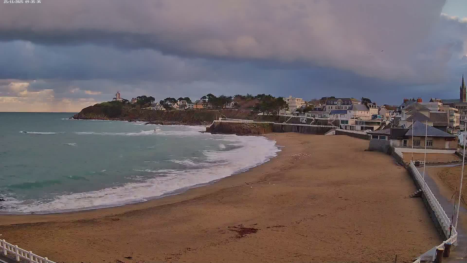 A wide sandy beach with breaking waves stretches towards a rocky headland topped by a lighthouse and houses, while a coastal town with numerous buildings and a prominent church spire lines the shore under a heavily overcast and moody sky.