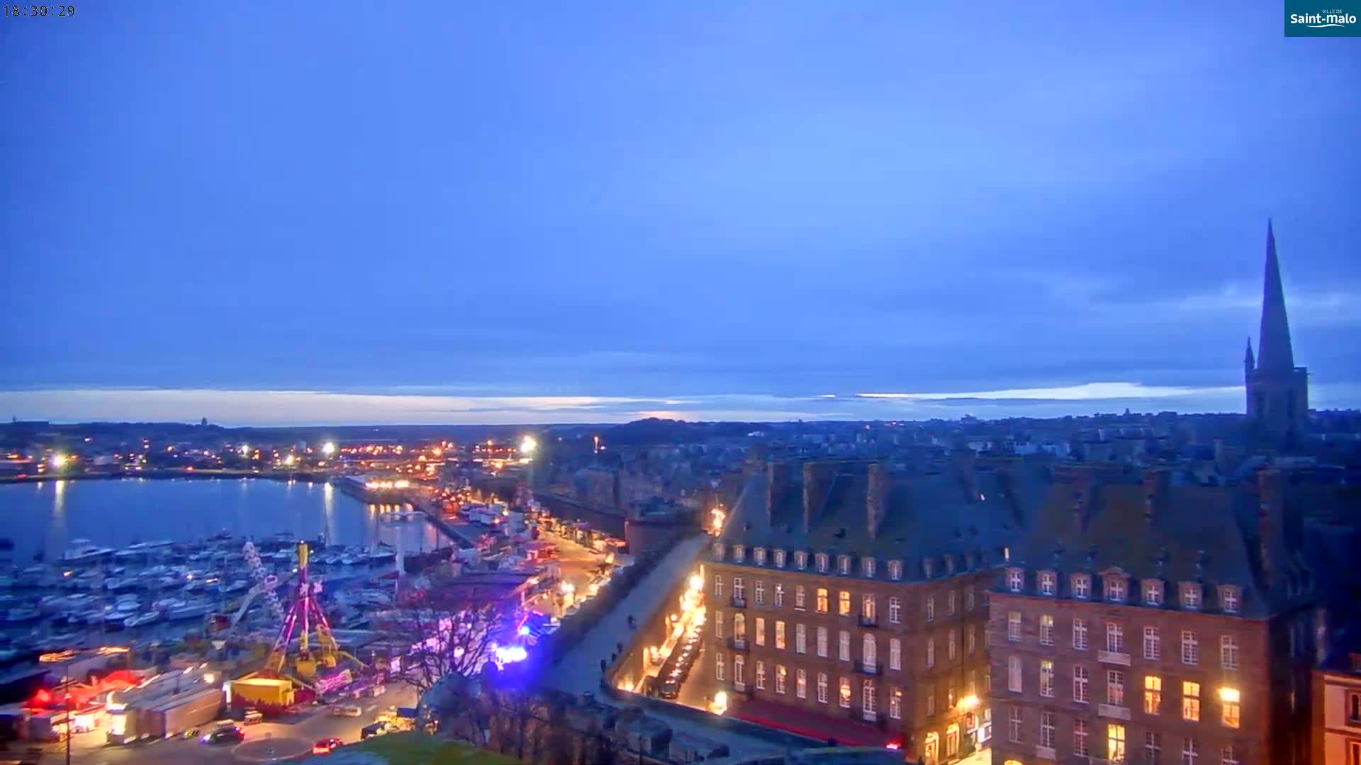 A twilight view of a harbor filled with boats, a waterfront city with illuminated buildings, and a tall church spire under a clear, dark blue sky.