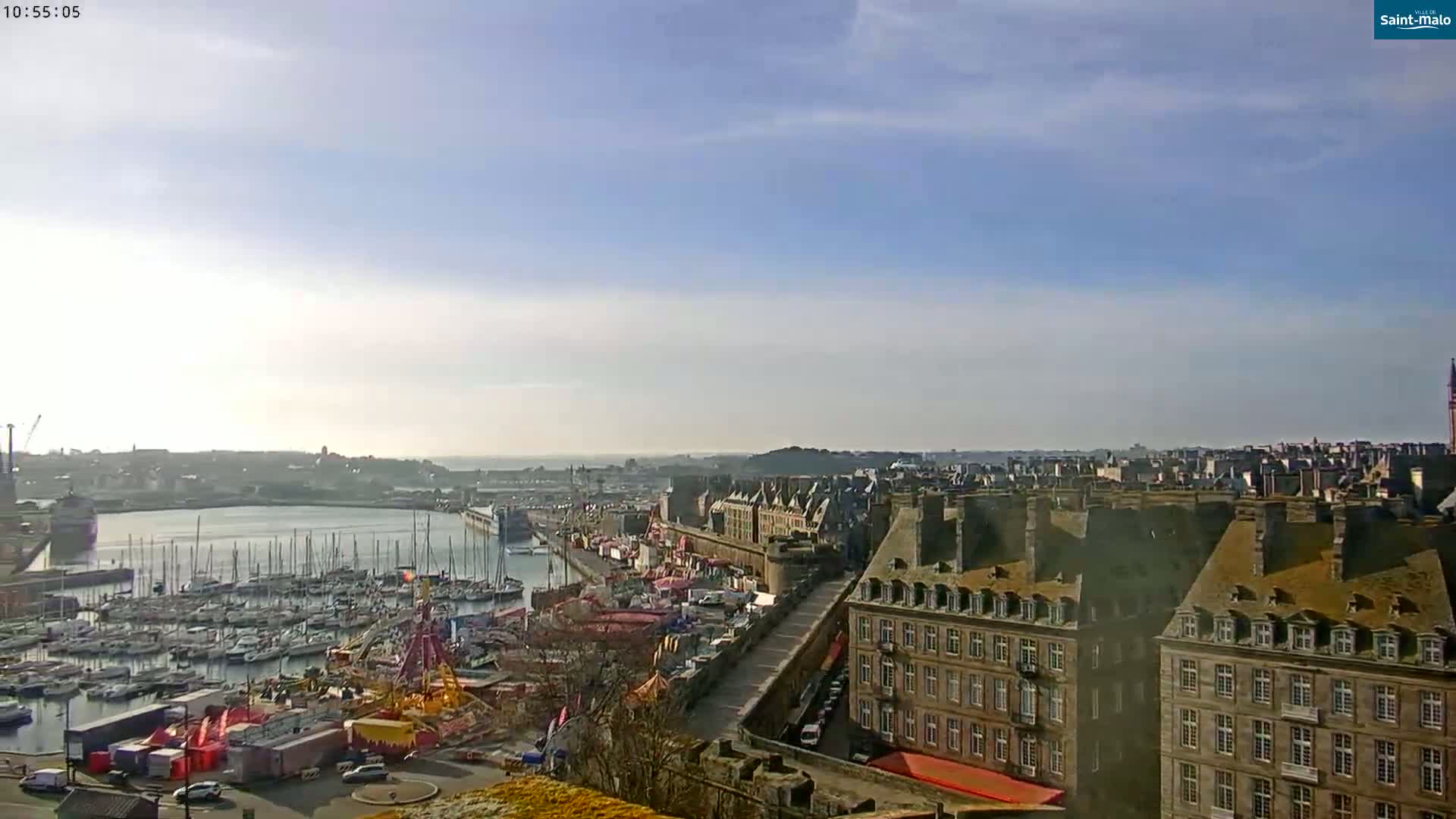 A high-angle, long shot shows a harbor filled with sailboats under a mostly sunny sky, next to a city with many buildings and a carnival-like atmosphere.