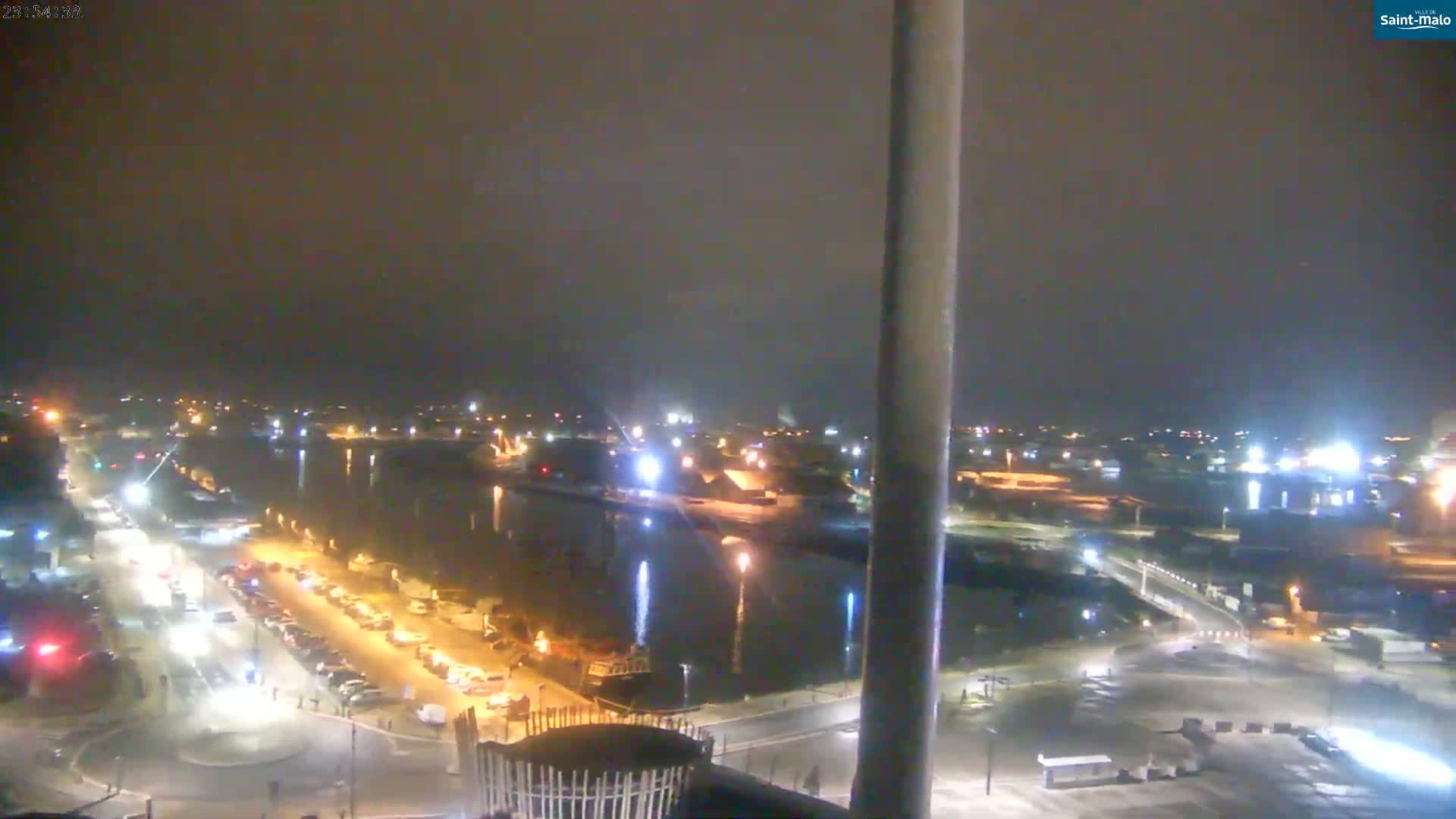 A nighttime aerial view shows a harbor with boats and cars parked along a road next to it.