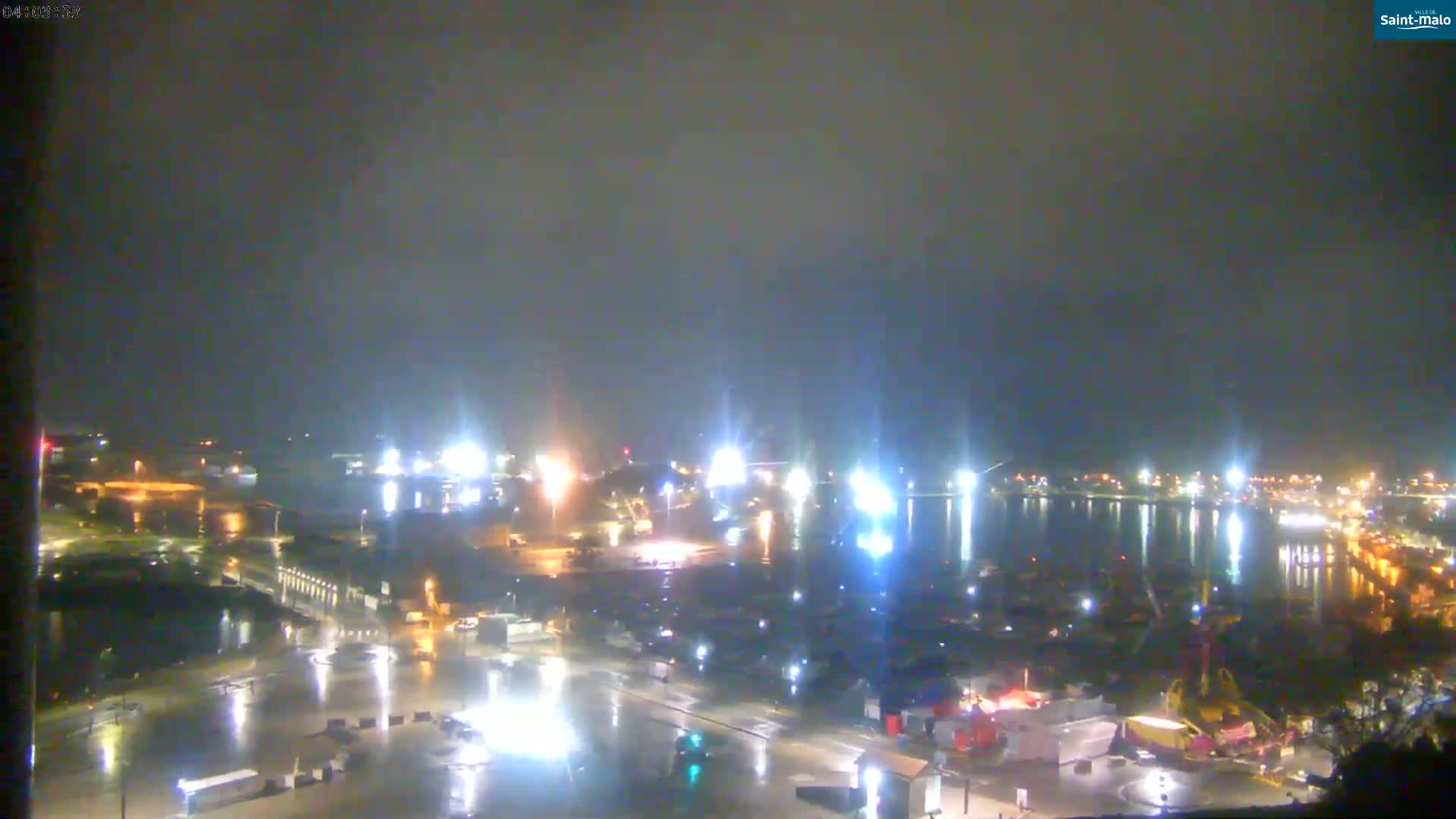 A nighttime view of a harbor area, illuminated by lights reflecting on wet streets and water under an overcast sky.