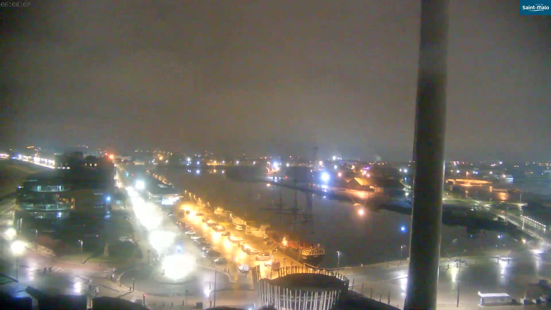 A nighttime, rain-soaked view of a harbor, showing a city waterfront with parked cars, buildings, and a large ship moored in the water.