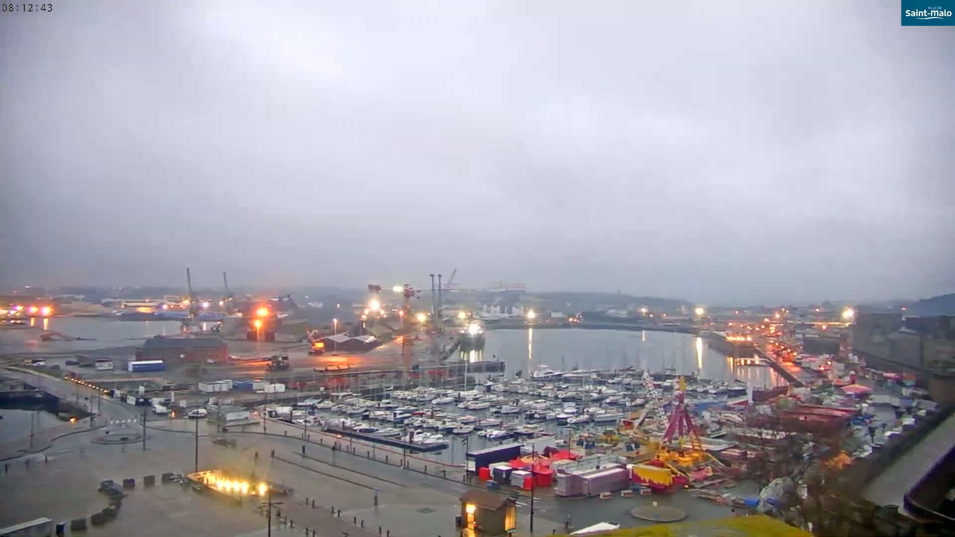 A harbor filled with numerous boats under an overcast sky, with a fairground visible nearby and a city in the background.