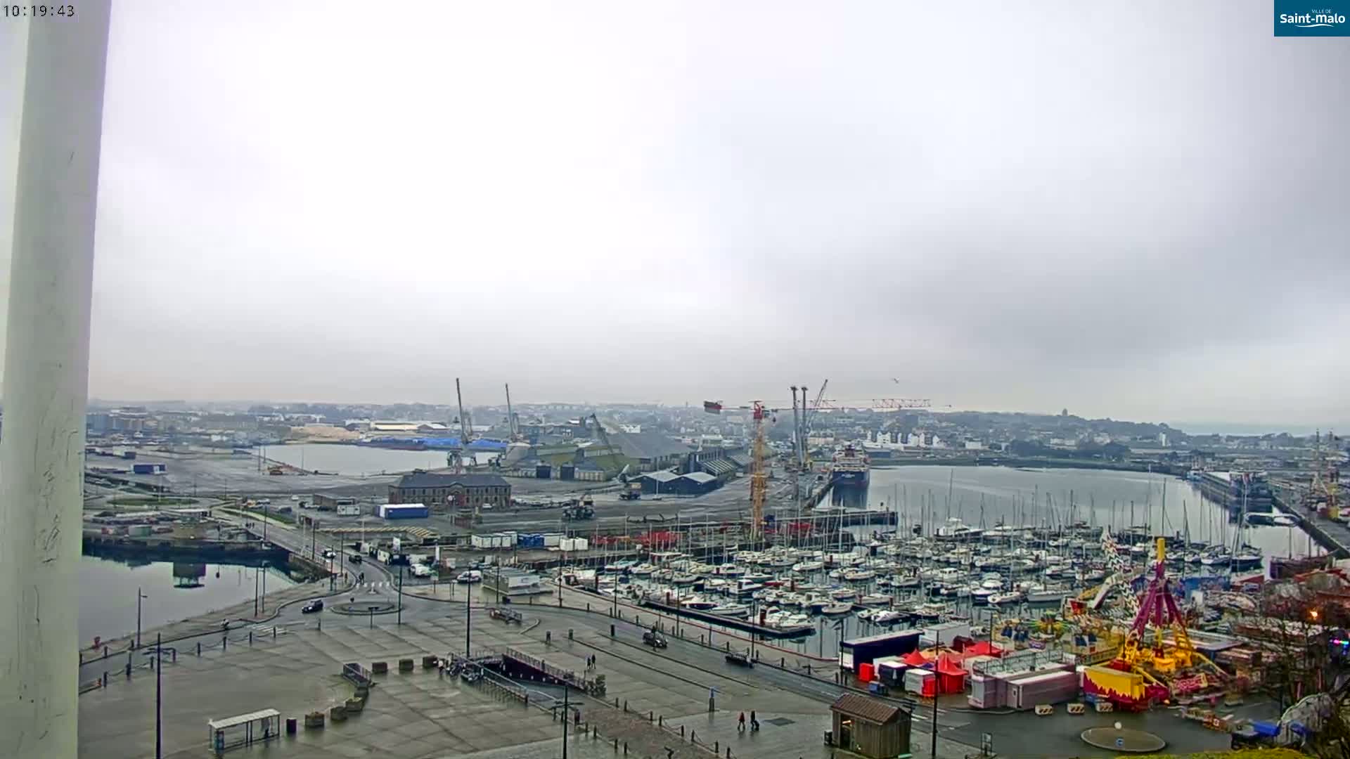A harbor filled with numerous boats under an overcast sky is shown, with cranes and industrial buildings visible in the background.