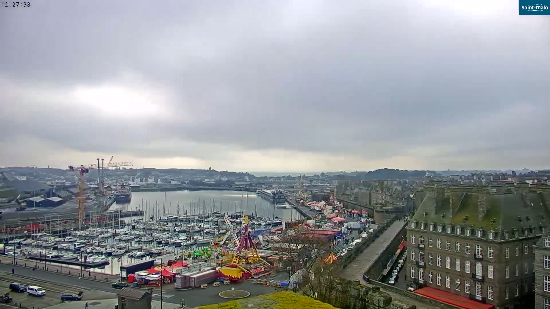 A harbor filled with sailboats is adjacent to a funfair, with a large building and overcast skies in the background.