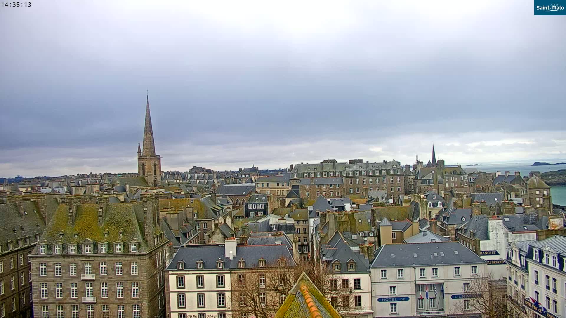 A sprawling cityscape of grey-roofed buildings under a cloudy sky, featuring a prominent church spire.