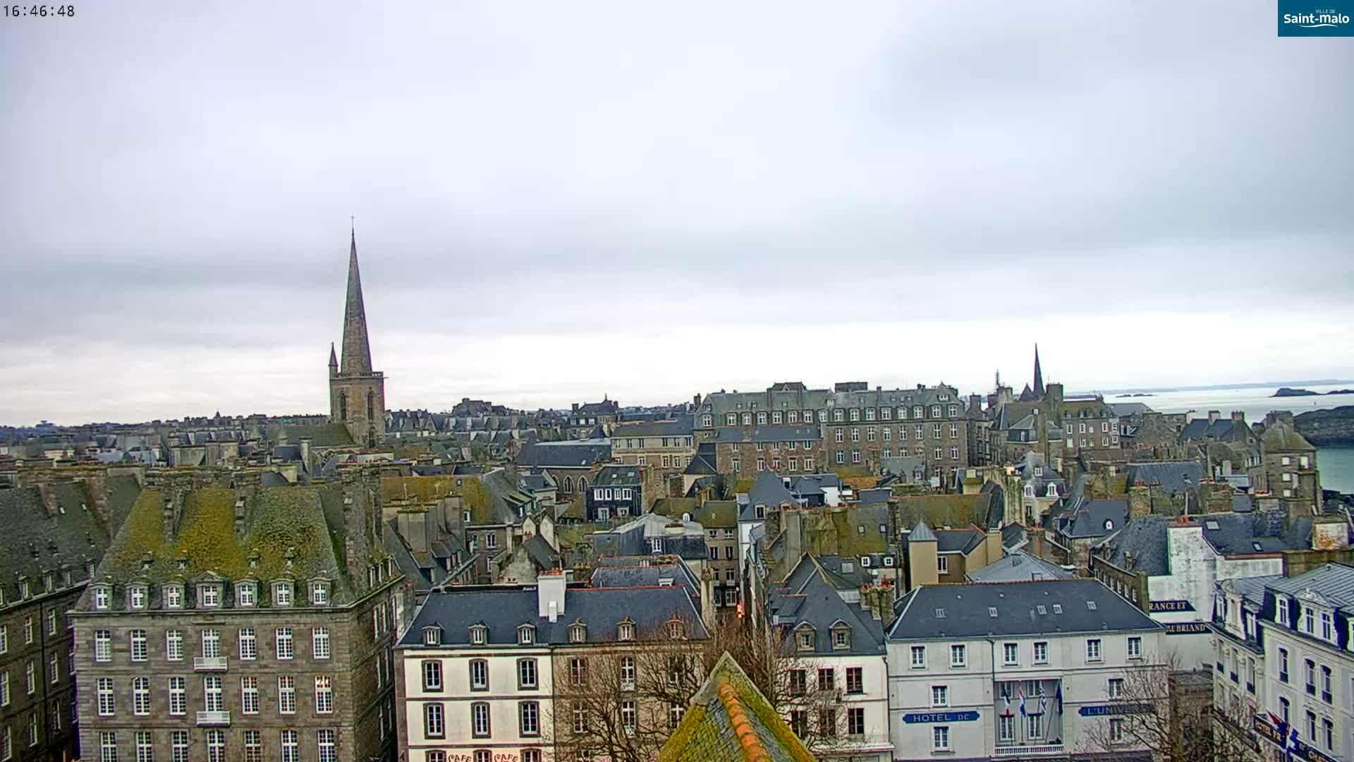 A high-angle, long shot of a city under an overcast sky shows a collection of buildings with dark slate roofs and a tall church steeple in the background.