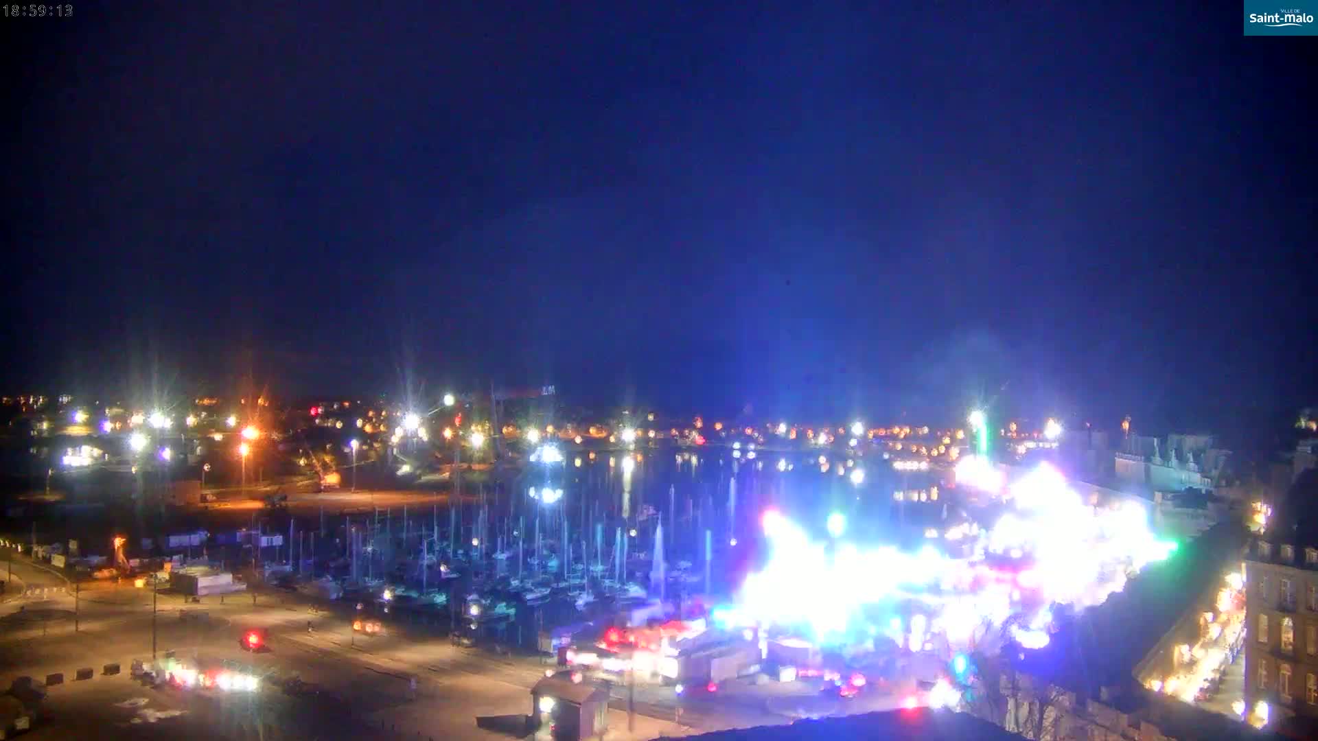 A brightly lit harbor at night, filled with boats, features a large illuminated spectacle in the foreground under a clear, dark blue sky.