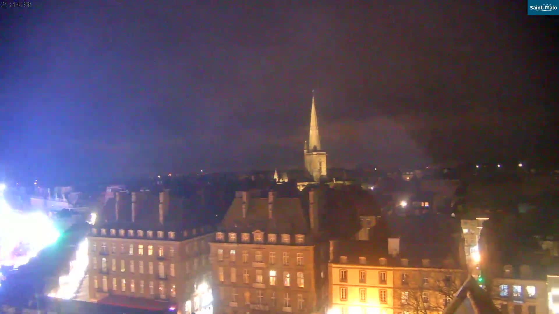 A nighttime, aerial view of a European city shows illuminated buildings and a tall church spire under a dark sky.