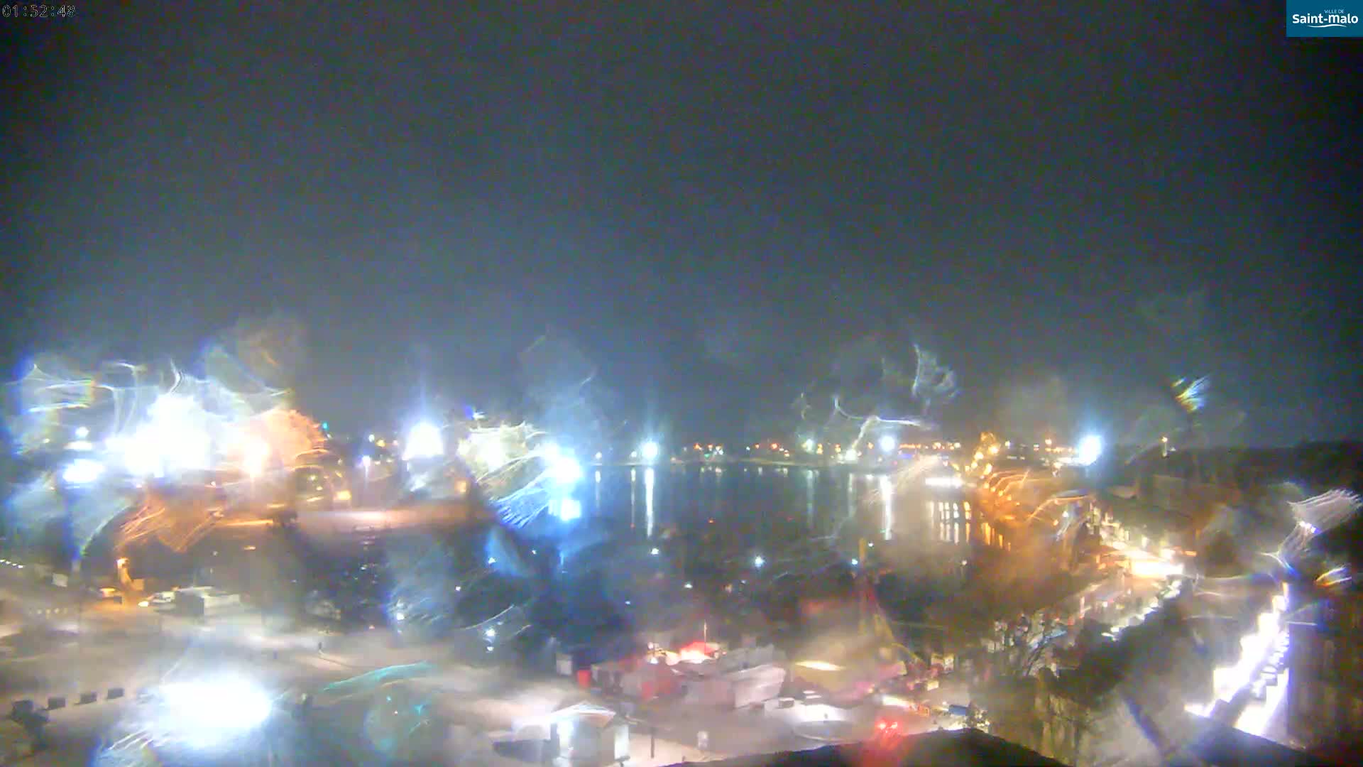 A nighttime view of a city waterfront with lights reflecting on the water, under a dark, possibly rainy or hazy sky.