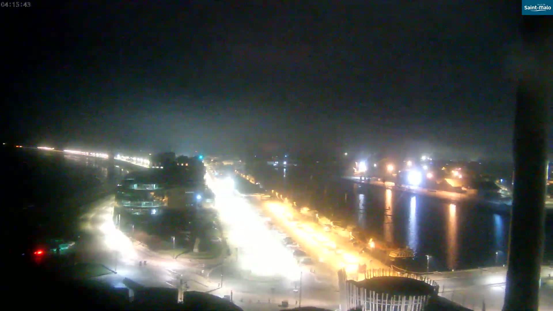 A nighttime, hazy view of a city waterfront with brightly lit streets and buildings, and a body of water reflecting lights.