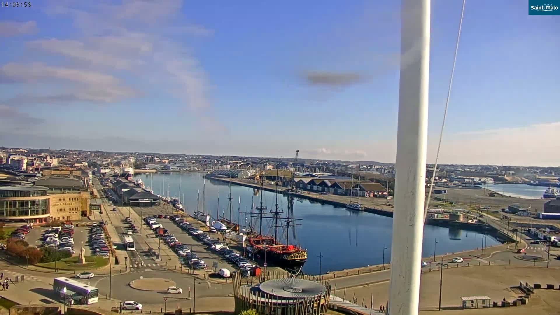 A mostly sunny harbor scene shows a large sailing ship docked near a city with numerous cars parked along a waterfront road.