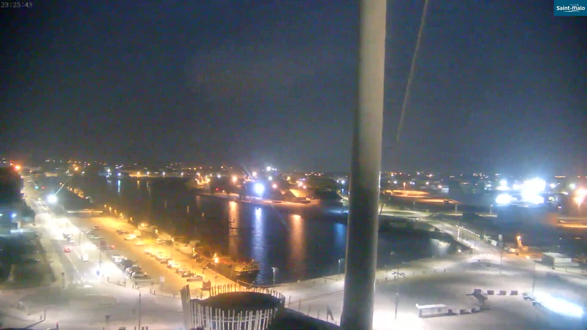 A nighttime, high-angle view of a harbor with a calm body of water reflecting lights from buildings and vehicles along a roadway.