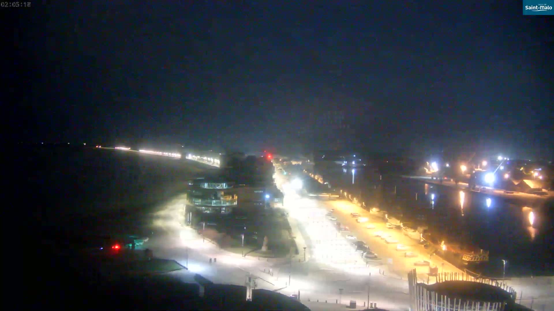 A nighttime, aerial view of a coastal town shows a dark beach, illuminated streets with parked cars, buildings, and a body of water with lights reflecting on its surface.