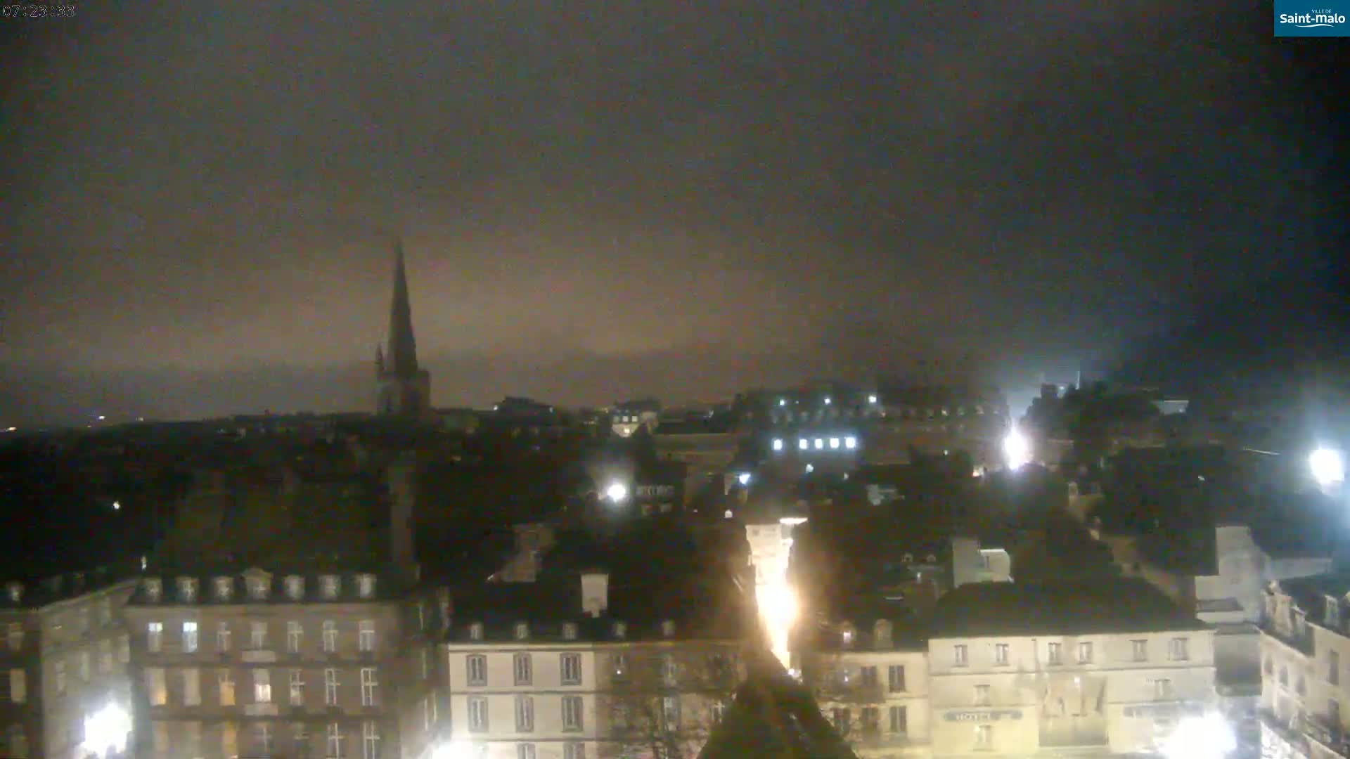 A nighttime, hazy view of a city with numerous buildings, including a tall church spire, illuminated by scattered lights.
