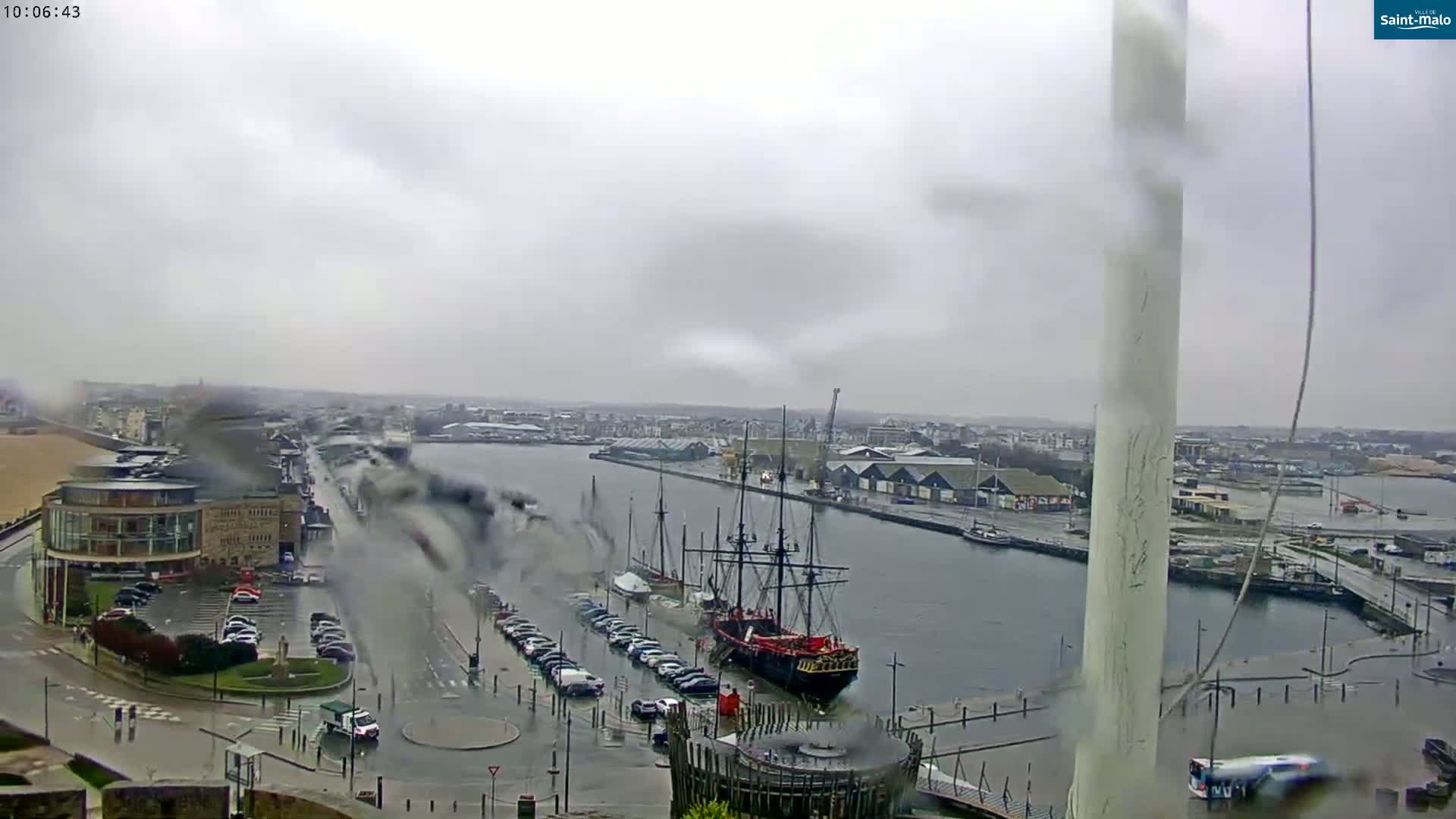 A rainy view of a harbor with a large docked ship, surrounding buildings, and parked cars.