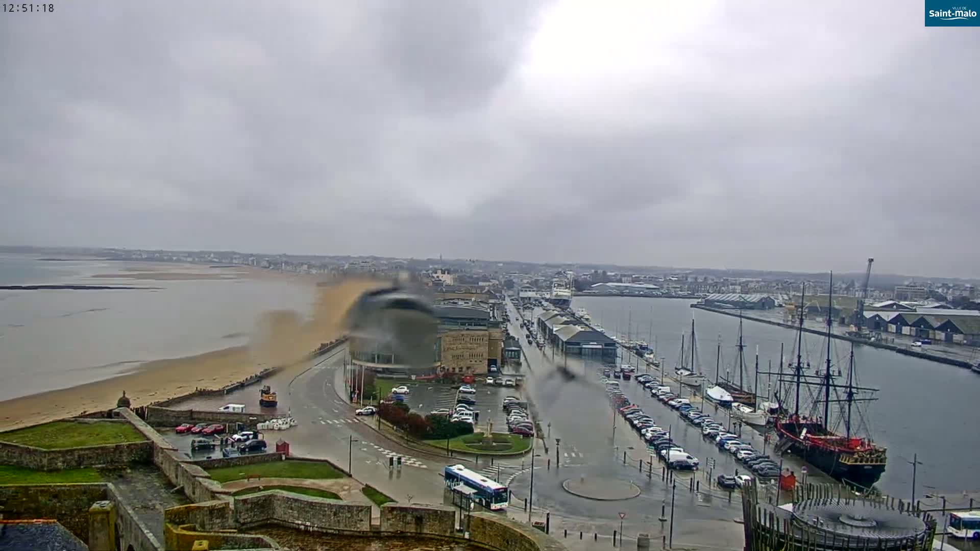 A rain-swept coastal city with a harbor full of boats, a sandy beach, and a large cloud of sand or dust rising near the shore.
