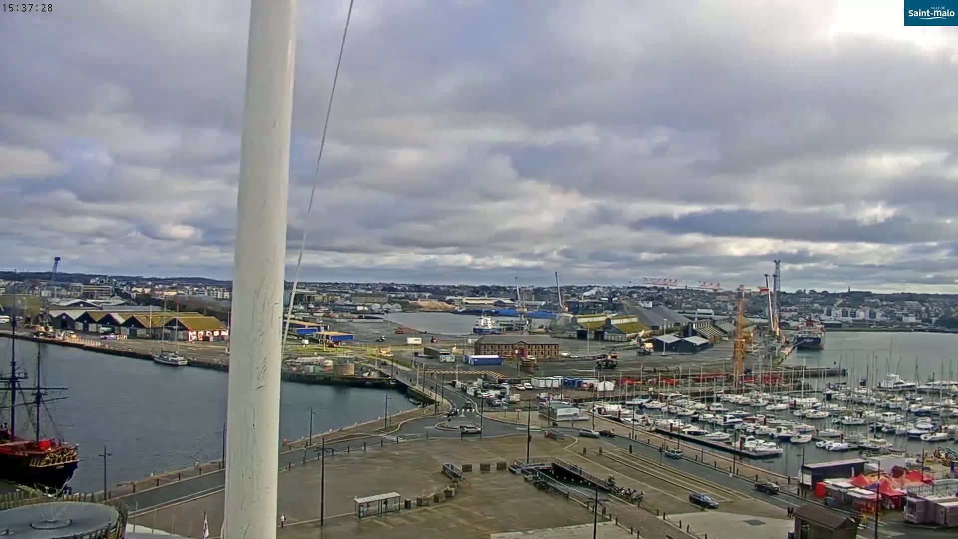 A harbor with numerous boats and ships is seen under a cloudy sky, with a large city in the background.