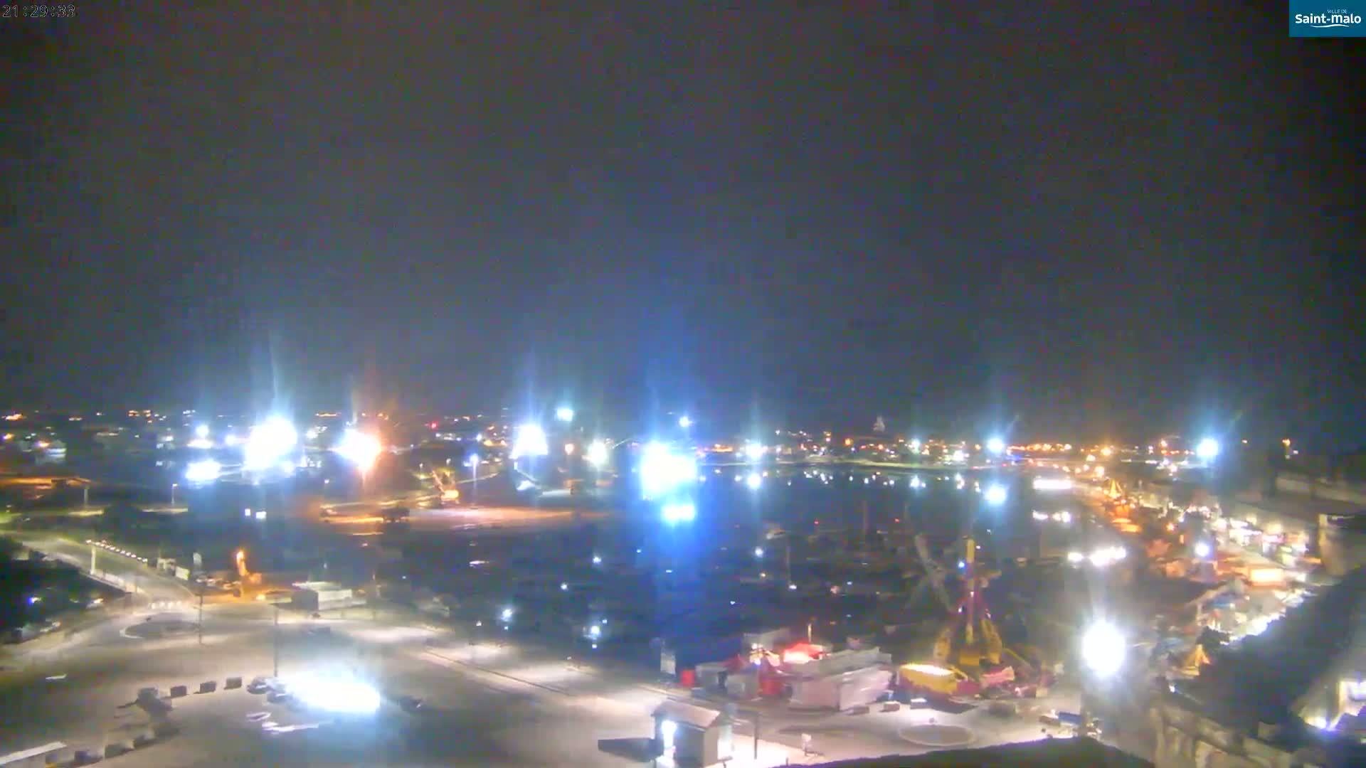 A nighttime, brightly lit harbor scene with numerous boats and a fairground visible, under a clear, dark sky.