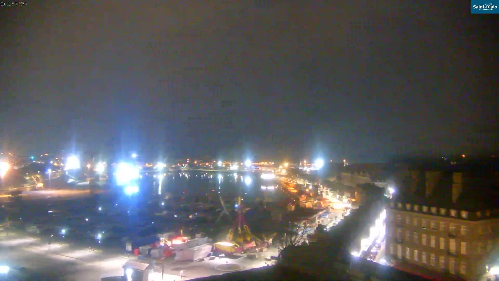 A nighttime, aerial view shows a harbor with boats, a waterfront area with buildings, and a mostly clear sky.