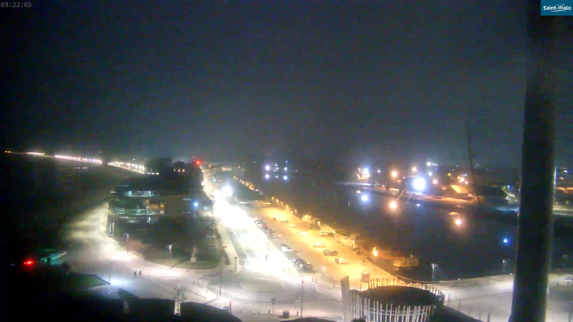 A nighttime, aerial view shows a coastal road with cars, buildings, and a body of water, under hazy conditions.