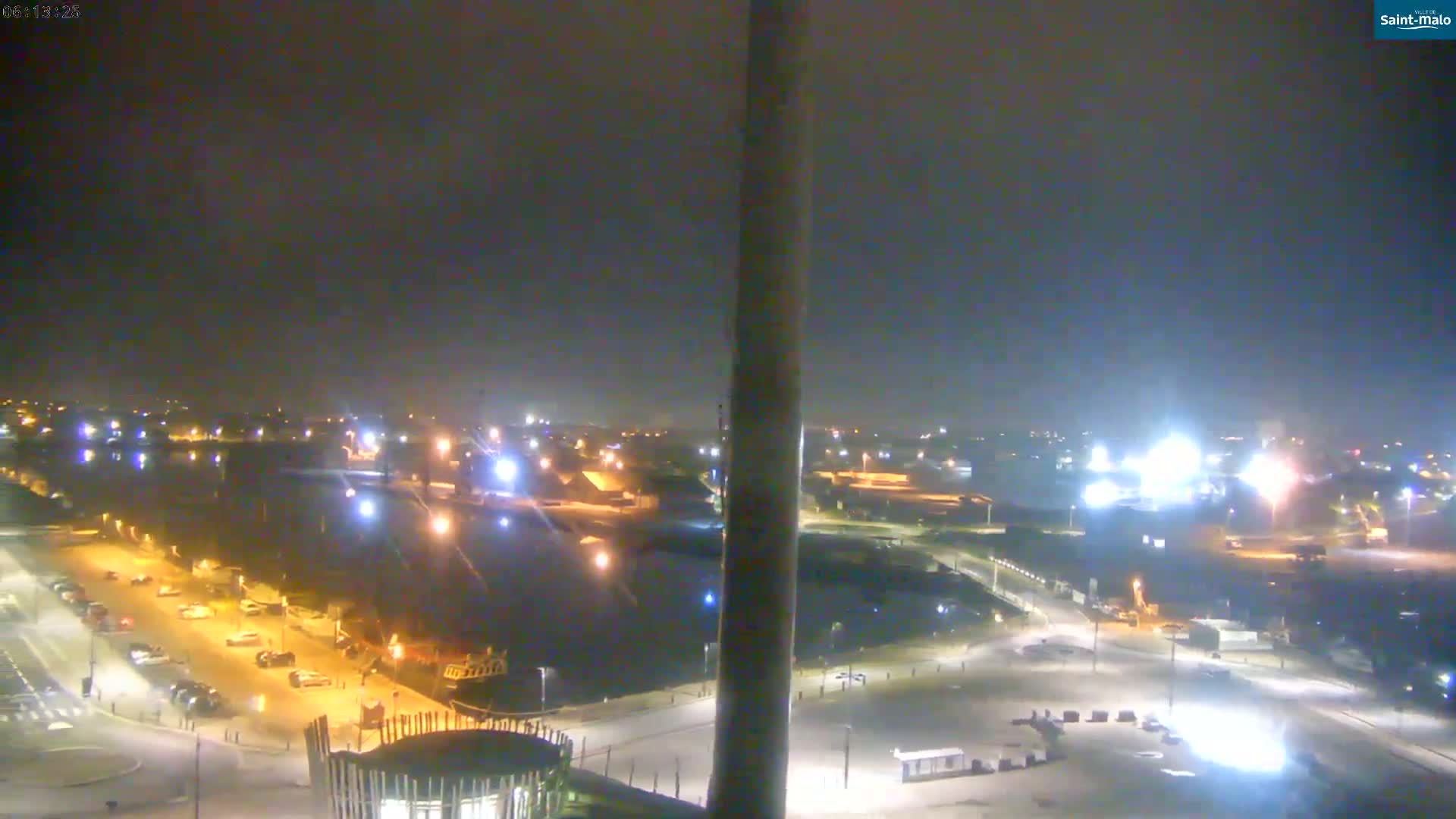 A nighttime, hazy view of a port area with many lights illuminating parked cars and buildings.