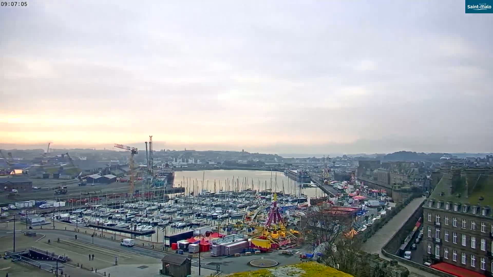 A sunrise over a harbor filled with sailboats and a fairground, under a mostly cloudy sky.