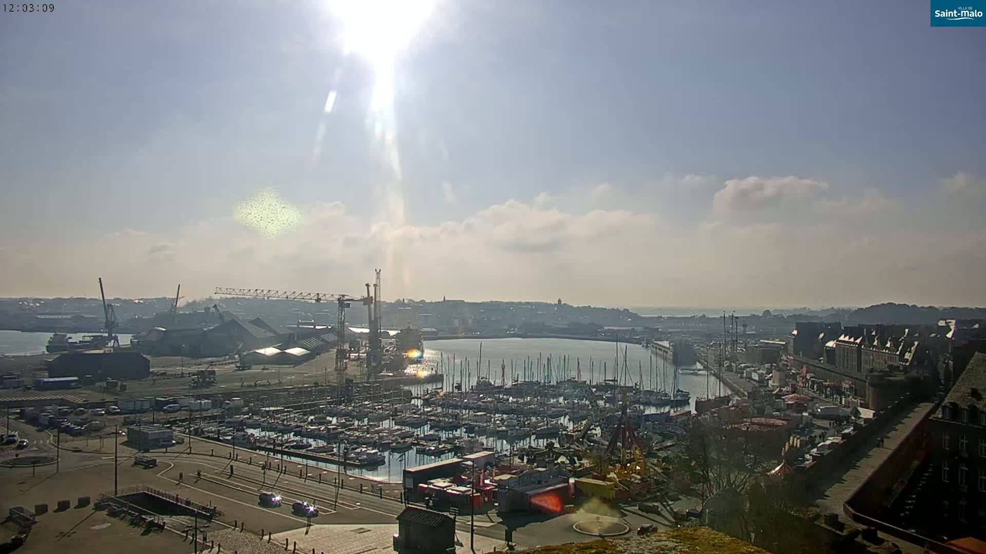 A sunny day overlooking a harbor filled with numerous boats and a cityscape beyond, with construction cranes visible near the docks.
