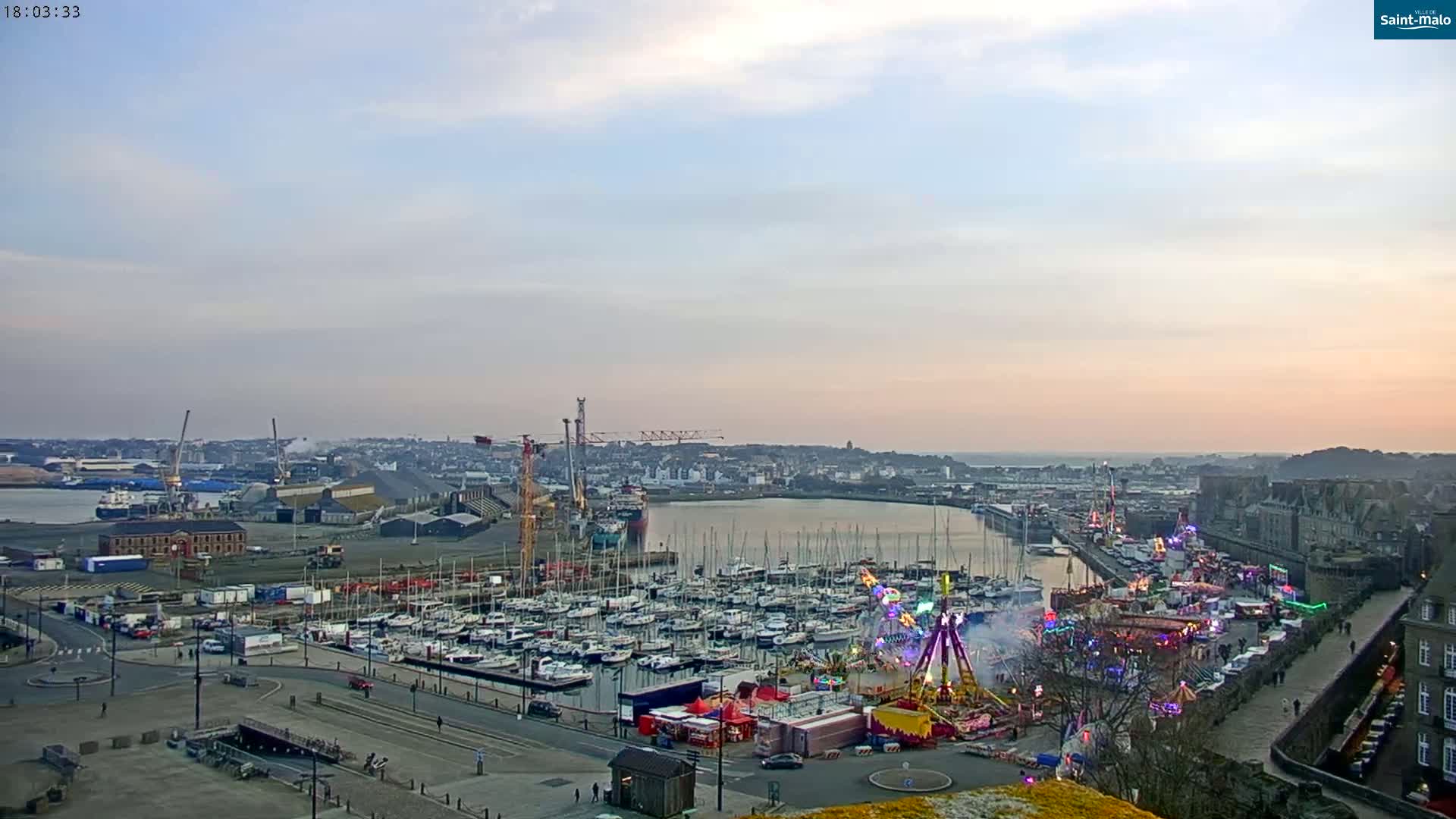 A harbor filled with sailboats at sunset, next to a fairground with carnival rides, under a mostly clear sky.