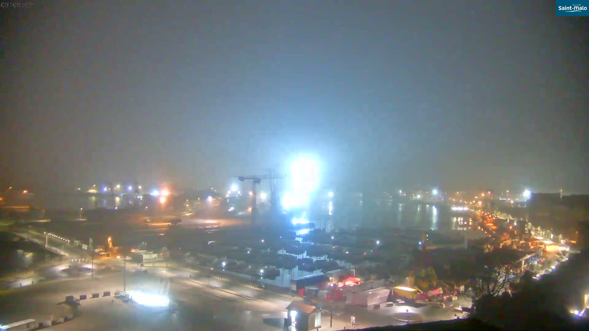 A foggy night scene shows a brightly lit construction site near a harbor with dimly lit buildings and roadways.