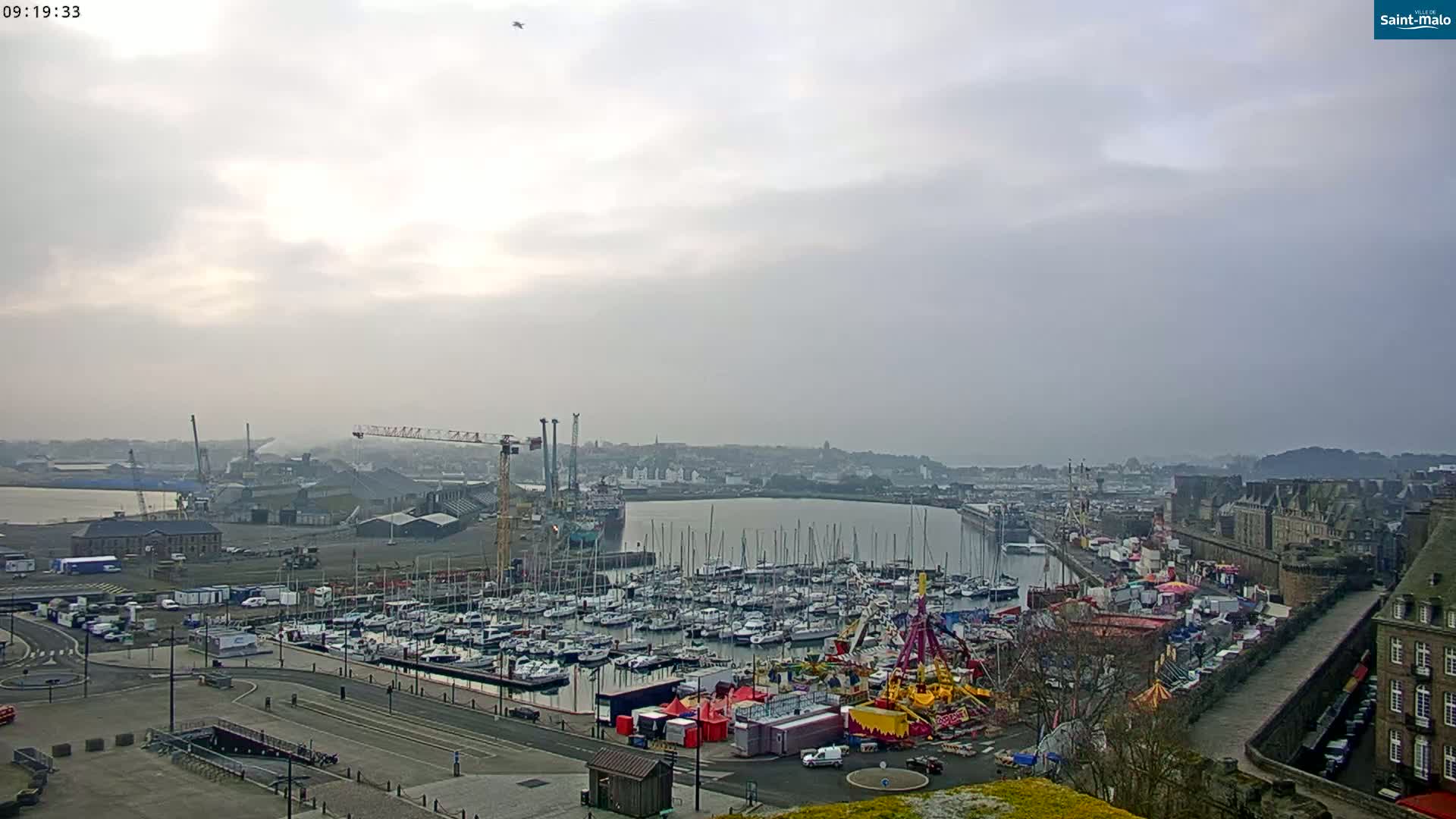 A hazy harbor scene shows a marina full of sailboats, a funfair, and a shipyard with cranes under a cloudy sky.
