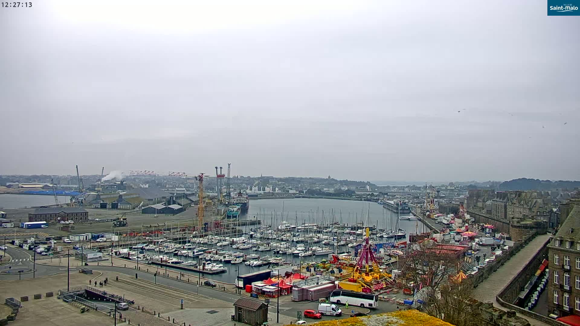 A harbor filled with numerous sailboats and a fairground is visible under an overcast sky.