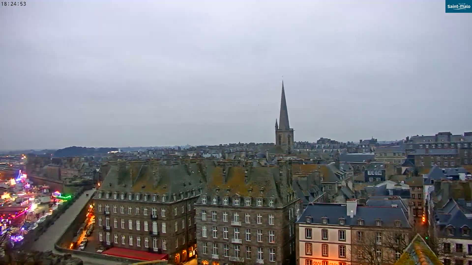 A high-angle, long shot of a city under an overcast sky shows many buildings with dark gray slate roofs, some lit up at night, and a tall church steeple in the distance.