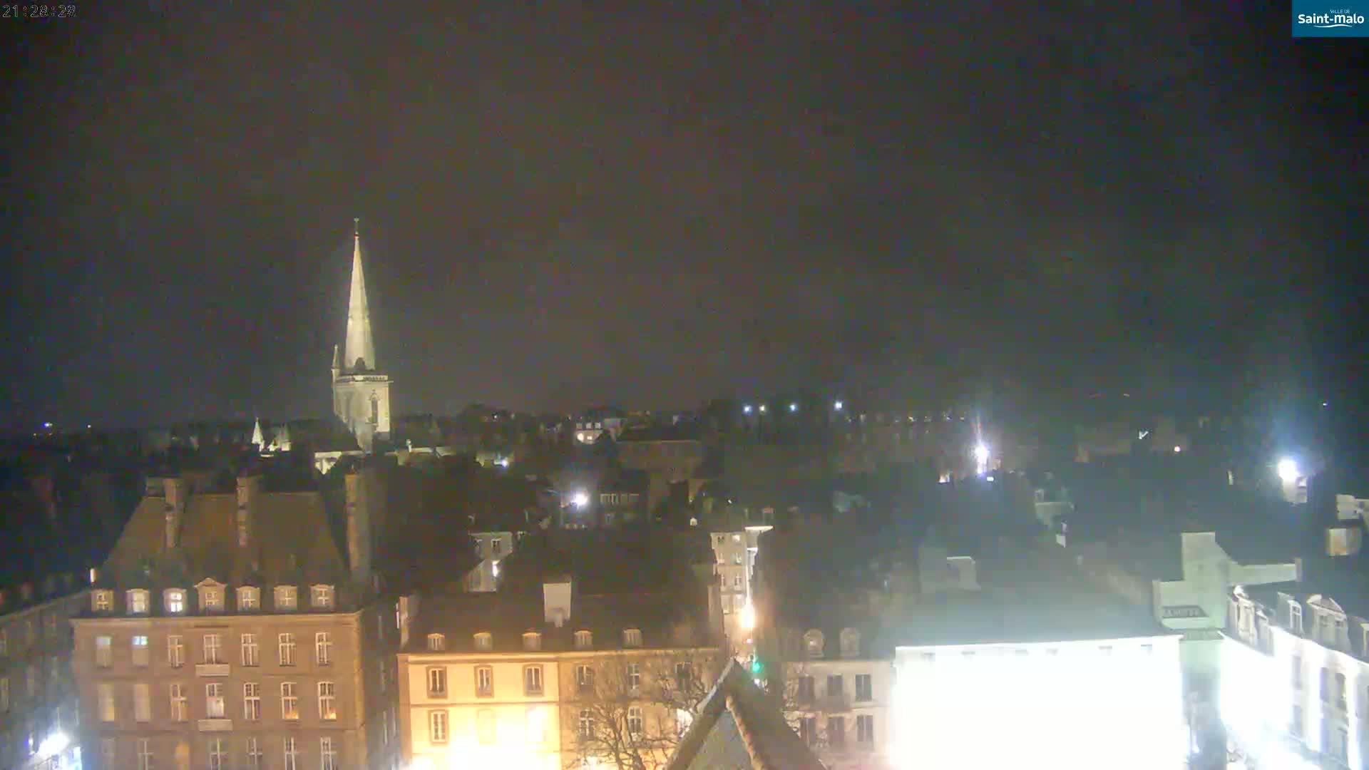 A nighttime, high-angle view of a city, featuring a tall church steeple and numerous buildings with illuminated windows under a dark, overcast sky.