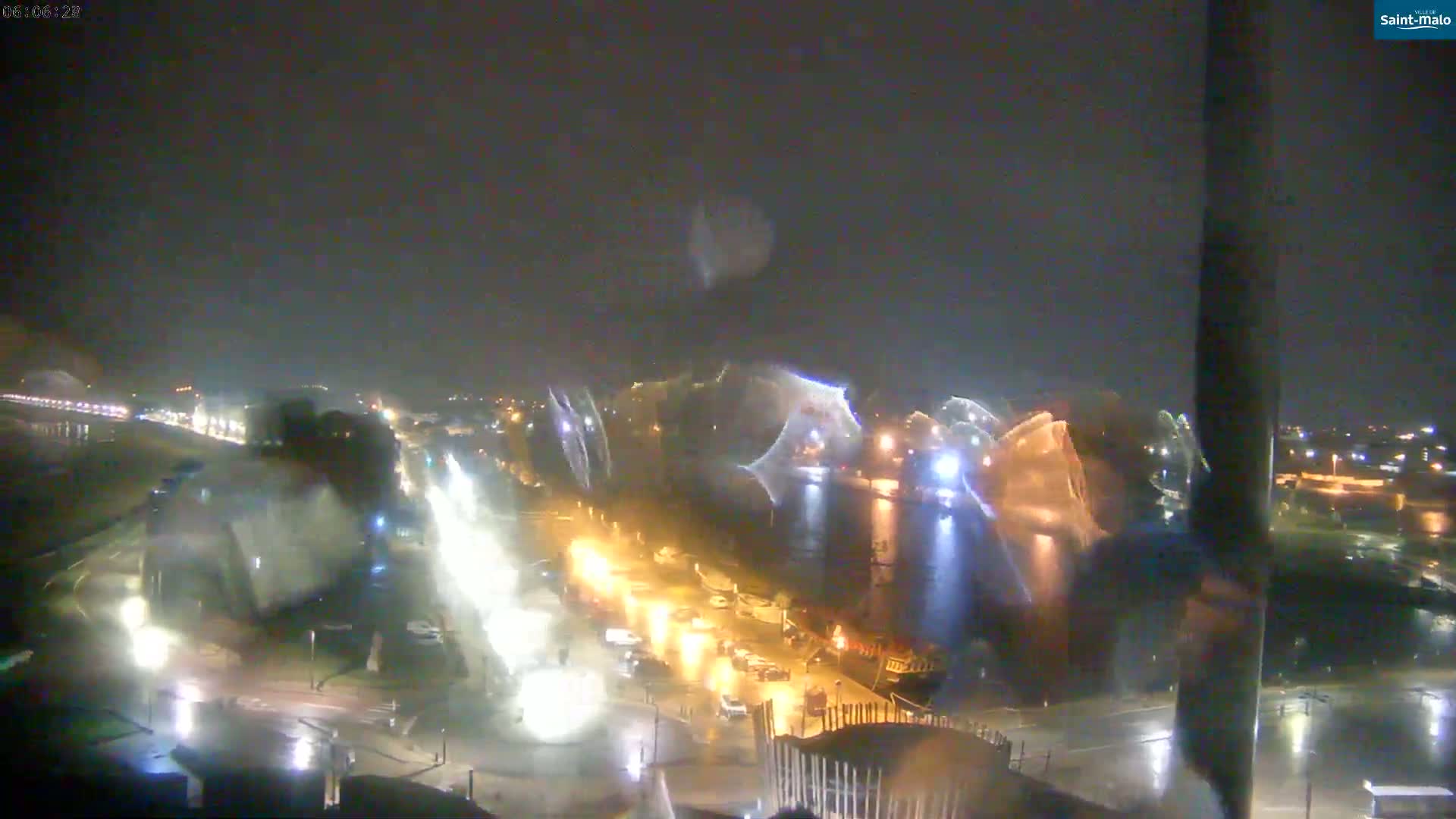 A nighttime view of a harbor with boats, cars, and lights, showing rain on a wet surface.