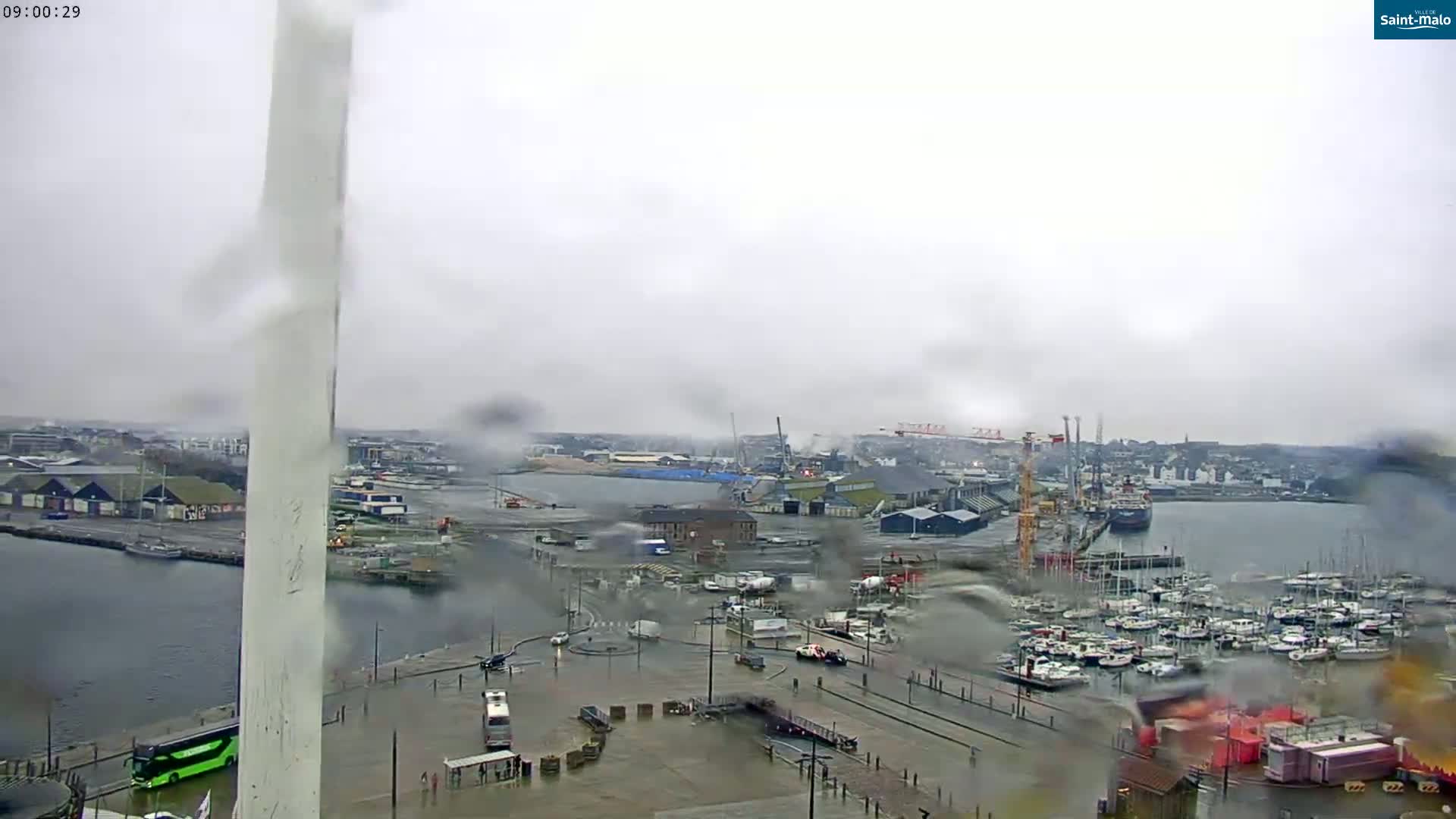 A rainy view of a harbor filled with numerous boats and ships, industrial buildings, and vehicles, seen from an elevated perspective.