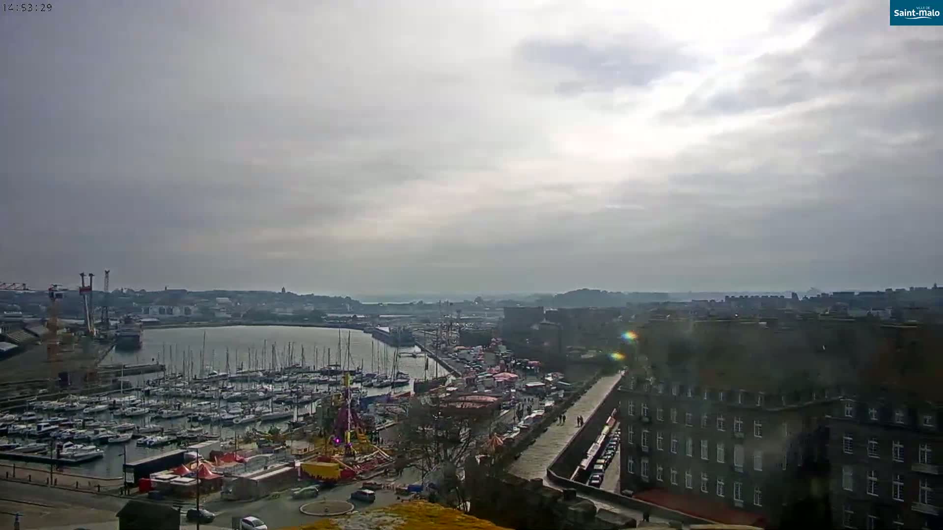 A harbor filled with sailboats is visible below a mostly cloudy sky, with a town and amusement park visible on the shore.