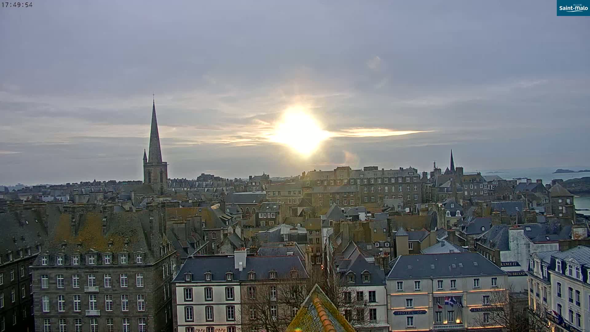 A mostly cloudy sunset illuminates a cityscape of closely packed buildings with several prominent church spires, overlooking a body of water.