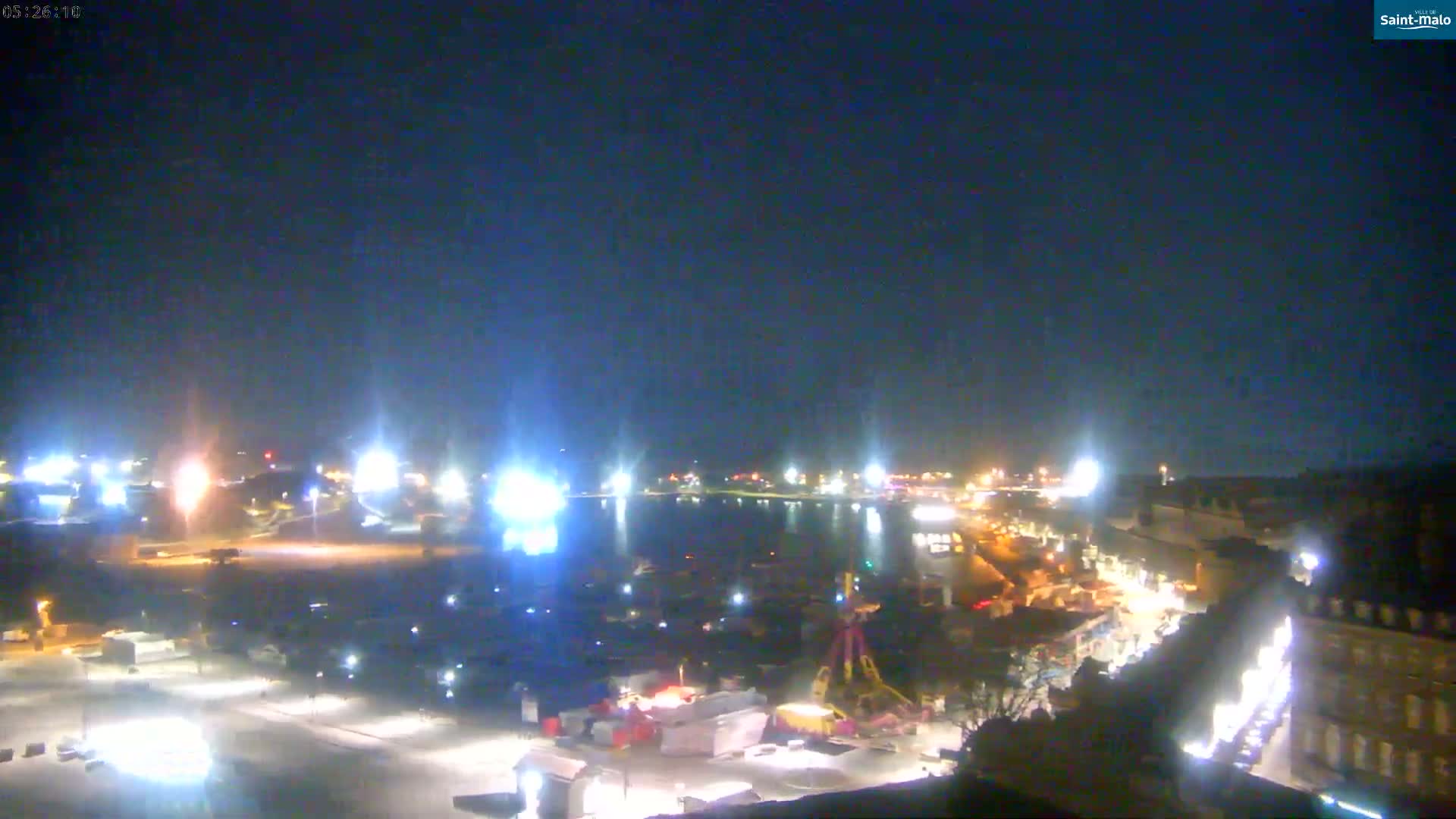 A nighttime aerial view of a harbor area with illuminated buildings and a fairground, under a clear dark sky.