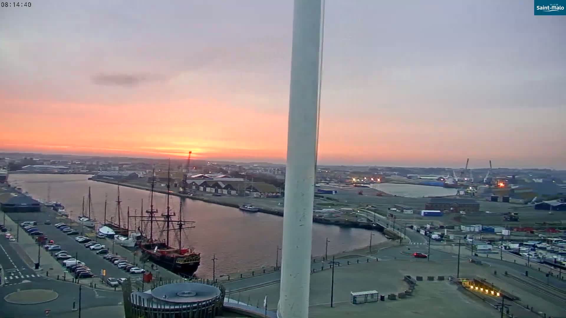 A sunrise over a harbor with docked ships, cars in a parking lot, and a tall white structure in the foreground.