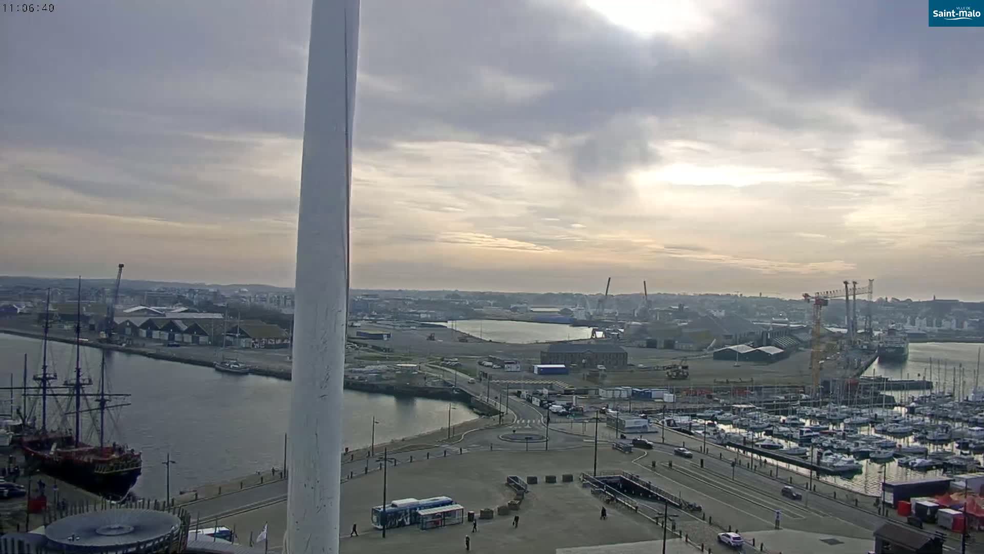 A mostly cloudy sky overlooks a harbor with docked sailboats, a large ship, and industrial areas.