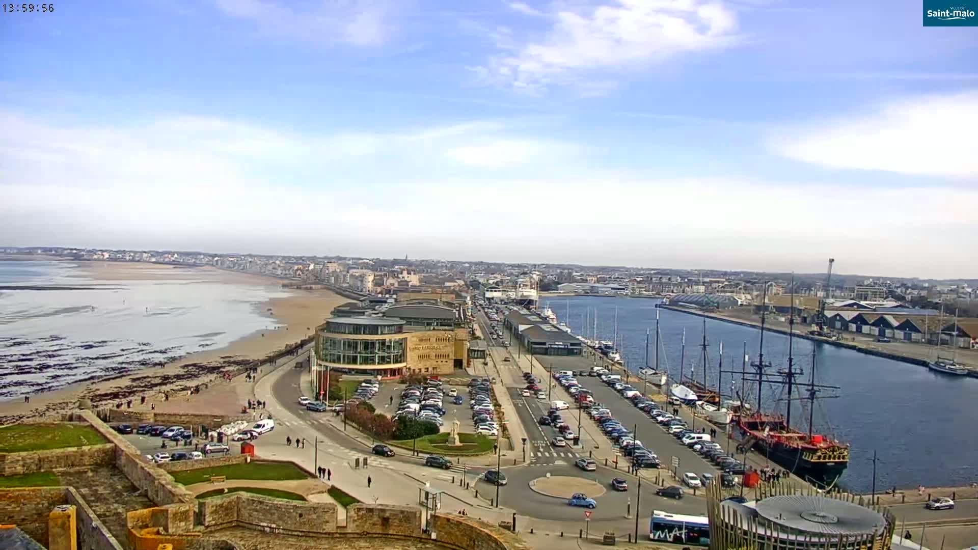 An aerial view shows a coastal city on a partly sunny day, with a sandy beach, harbor filled with boats, and a large building near the waterfront.
